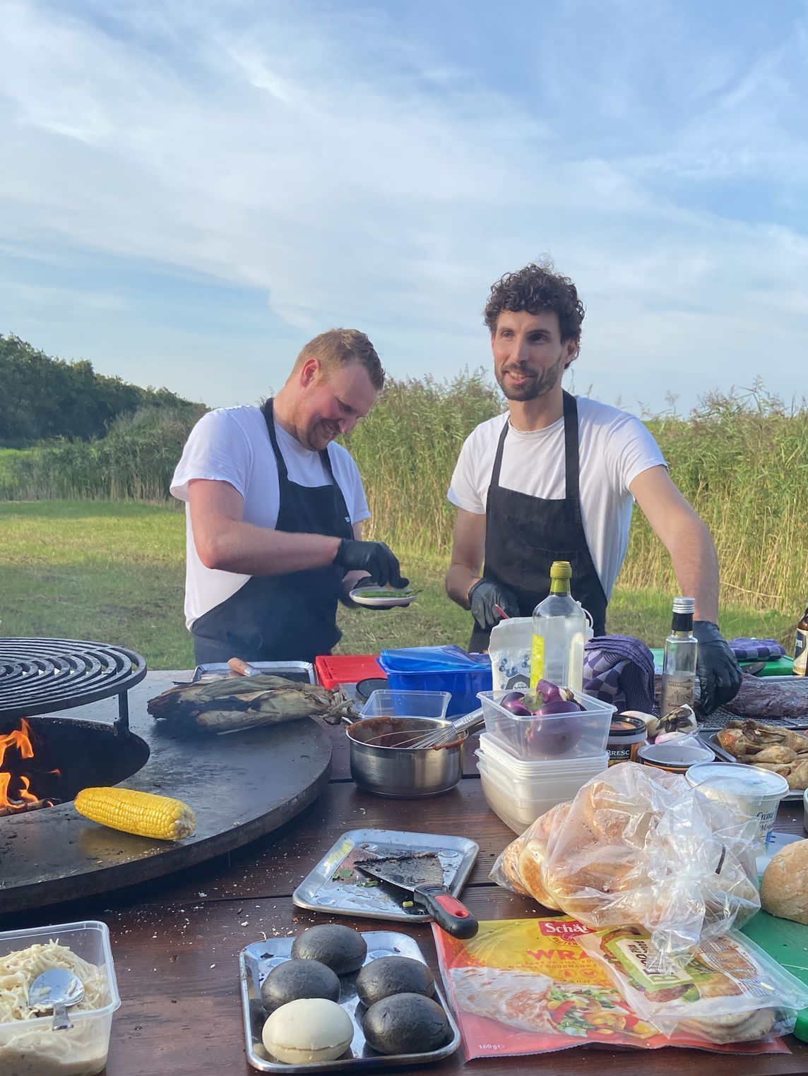 Twee mannen staan ​​buiten te barbecueën, beiden met schorten aan. De ene glimlacht, de andere kijkt toe. 