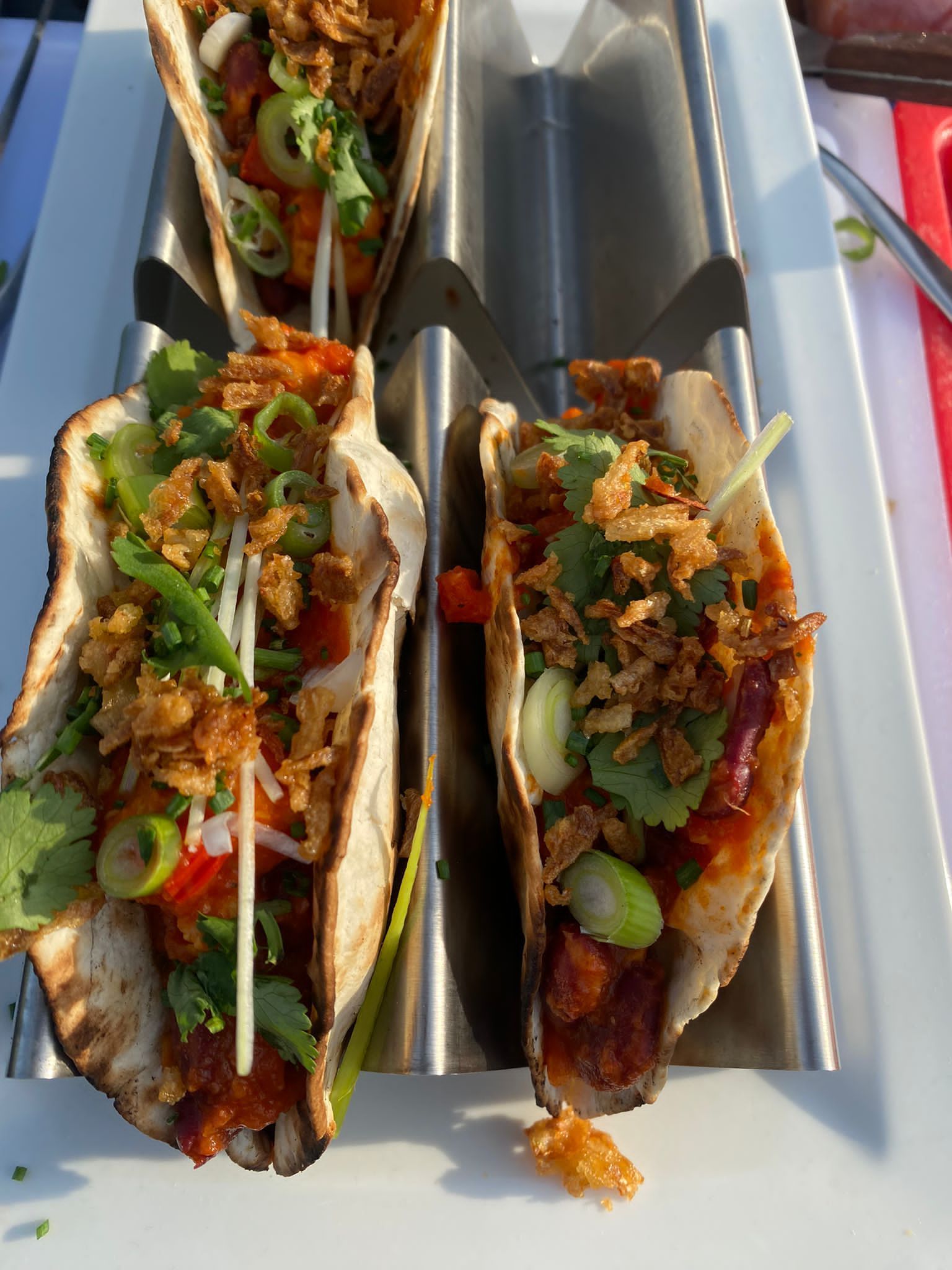 Drie gevulde taco's in zilverkleurige metalen houders, gegarneerd met lente-uitjes en gebakken uitjes.