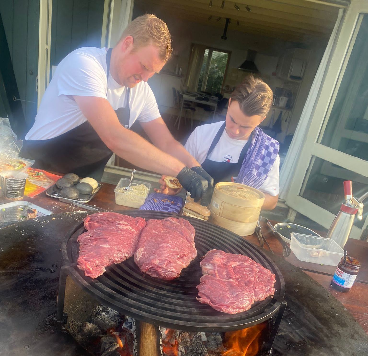 . Het vlees sist op de grill, een houten tafel is gedekt met eten.