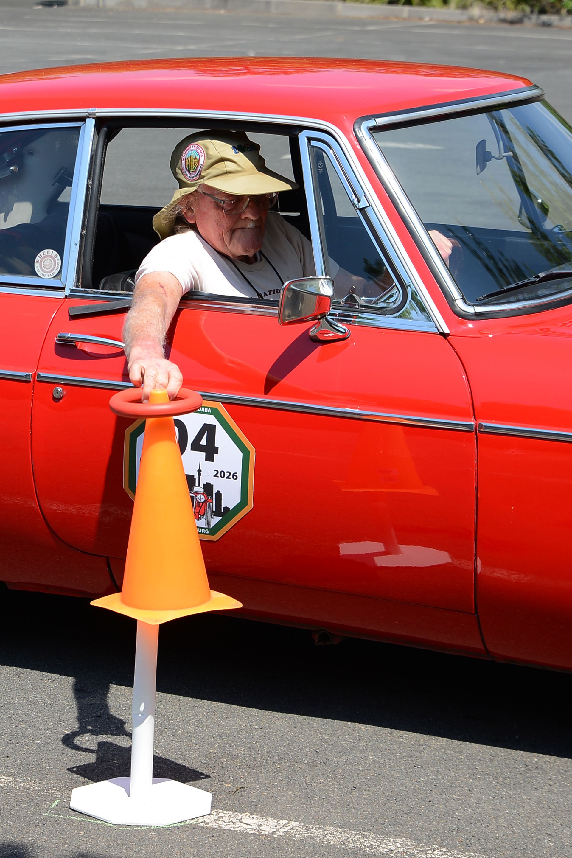 Person in a red car reaching toward an orange cone at a roadside checkpoint.