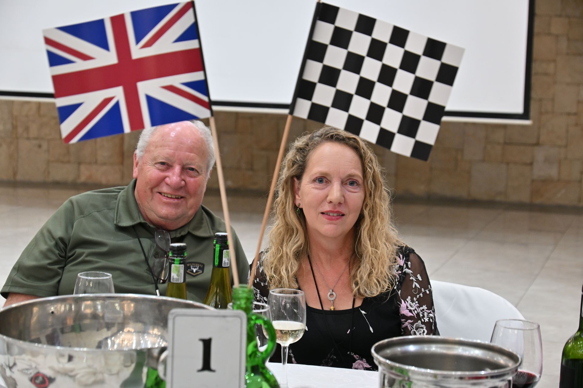 Two people seated at a table with UK and checkerboard flags behind them