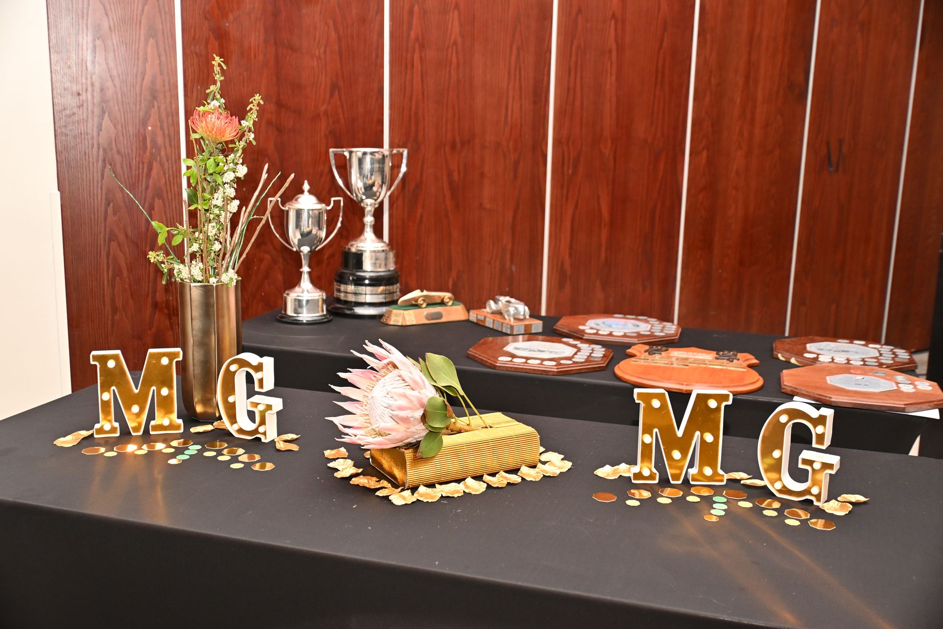 Gold “M.G” letters, silver trophies, and decorated treats on a black table against a wooden wall.