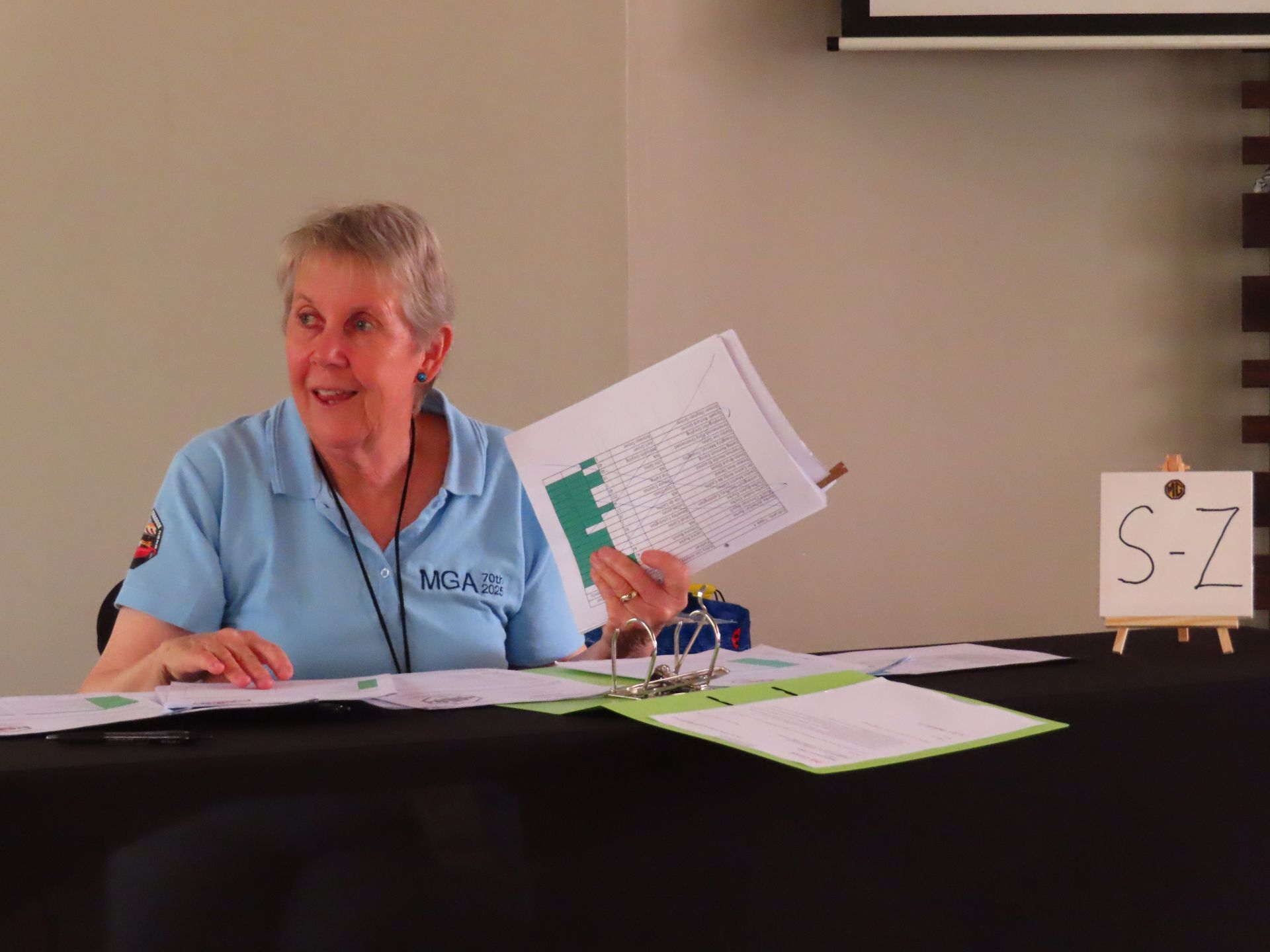 Woman at a table with papers, smiling and holding up a sheet in a light-blue shirt.