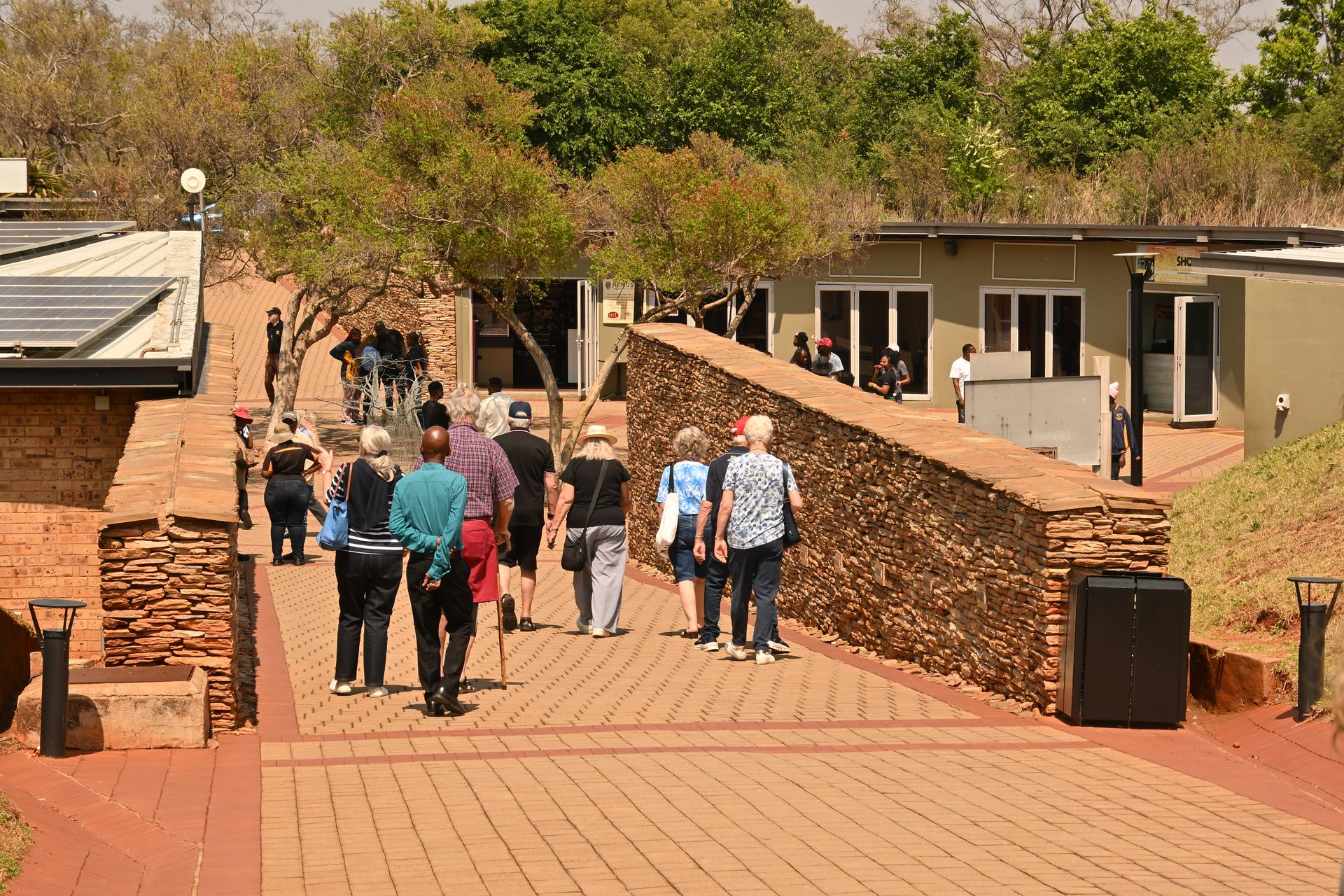 Group walking on a brick campus path between buildings and trees, with a brick wall and benches nearby.
