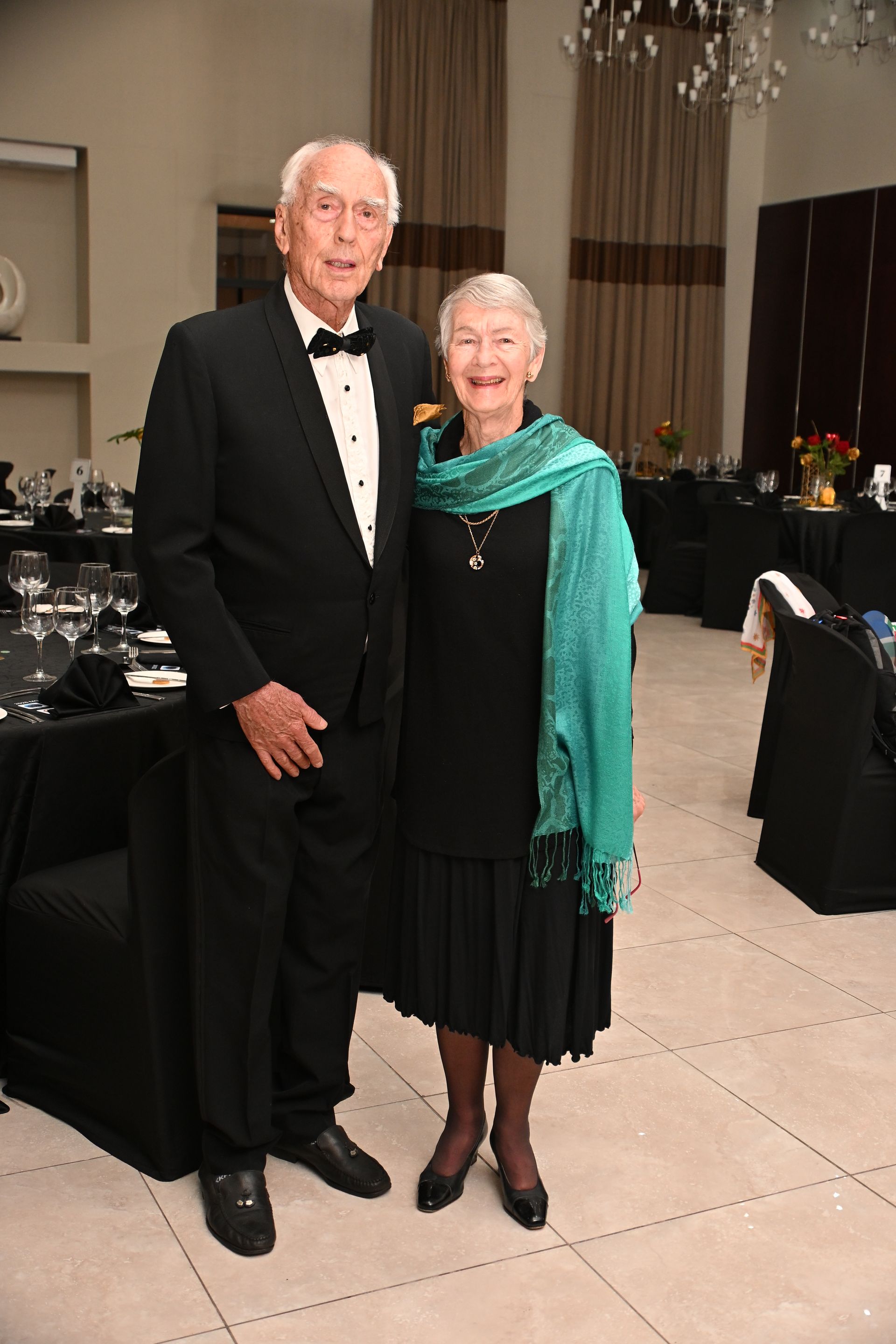 Two people in formal attire standing together at an indoor event, smiling near a table.