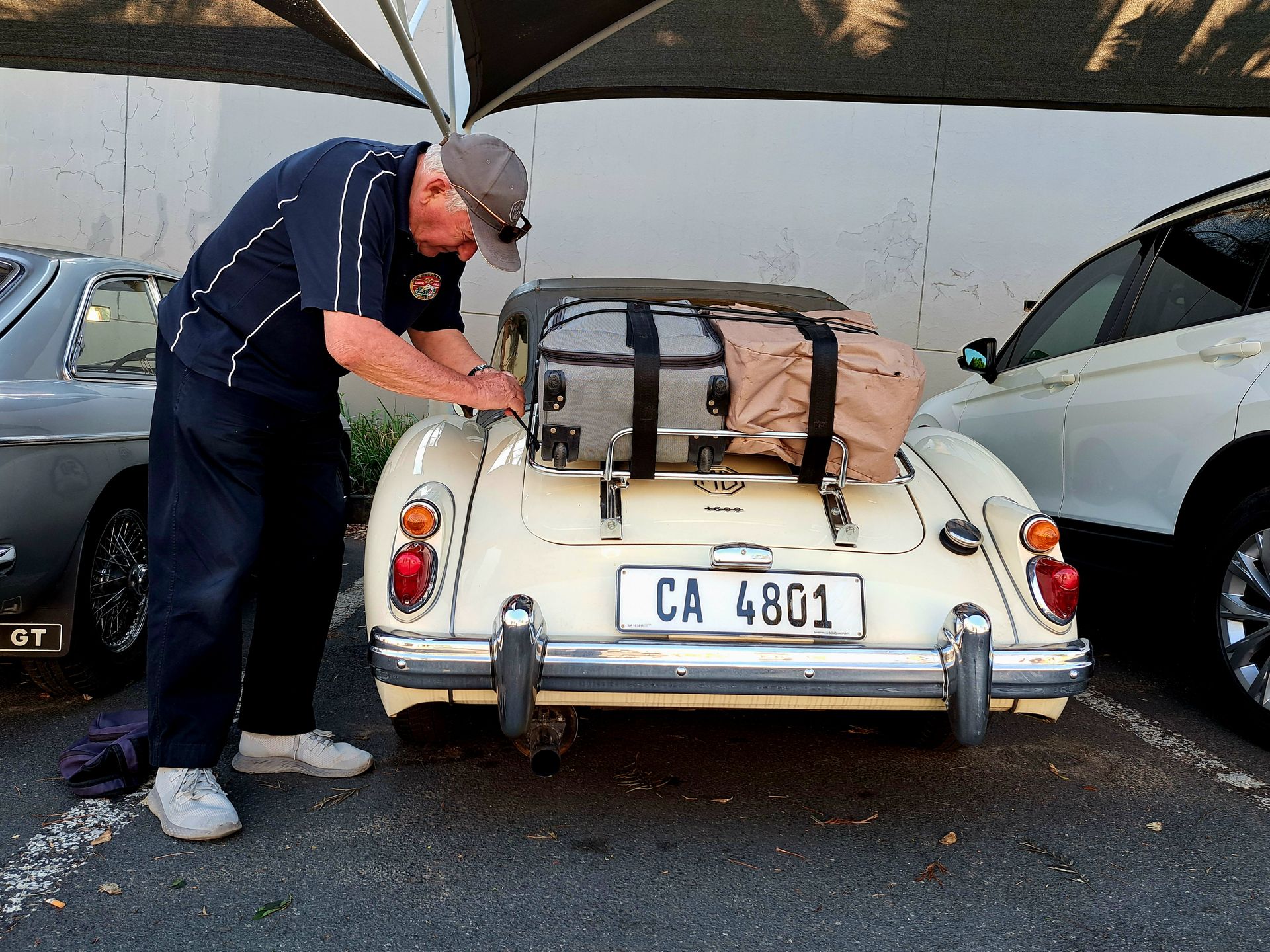 Man leaning into a cream vintage convertible parked between two cars, luggage strapped on the back.