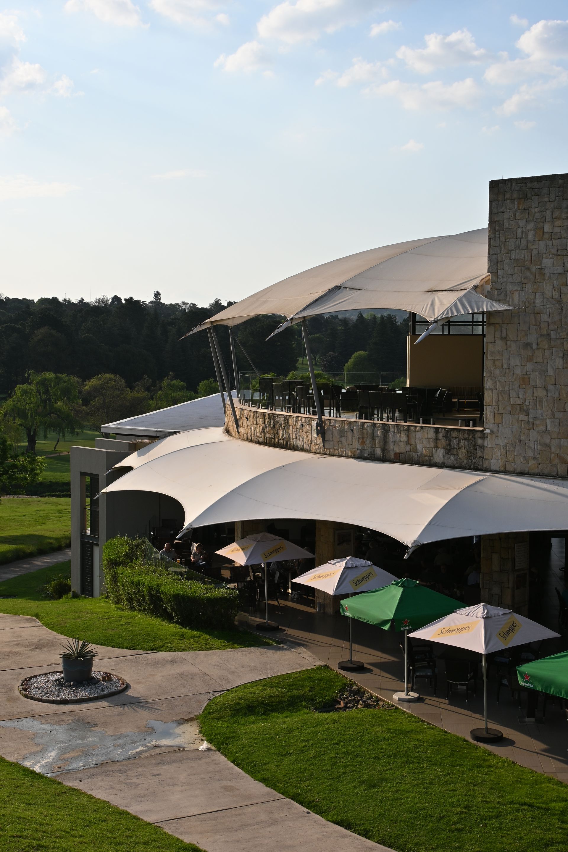 Outdoor resort terrace with white canopy umbrellas, green lawns, and a stone building under a partly cloudy sky