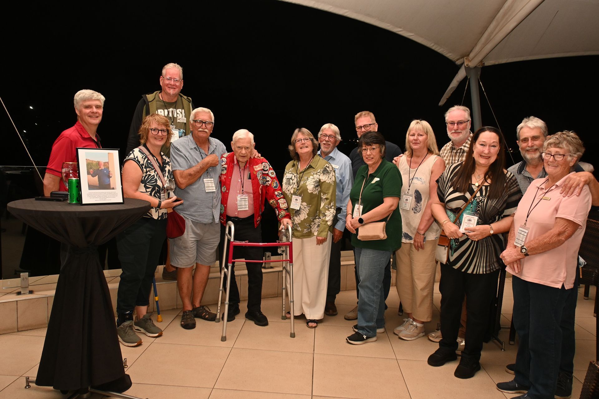 Group of people posing on a stage beside a framed award plaque under a tent canopy.