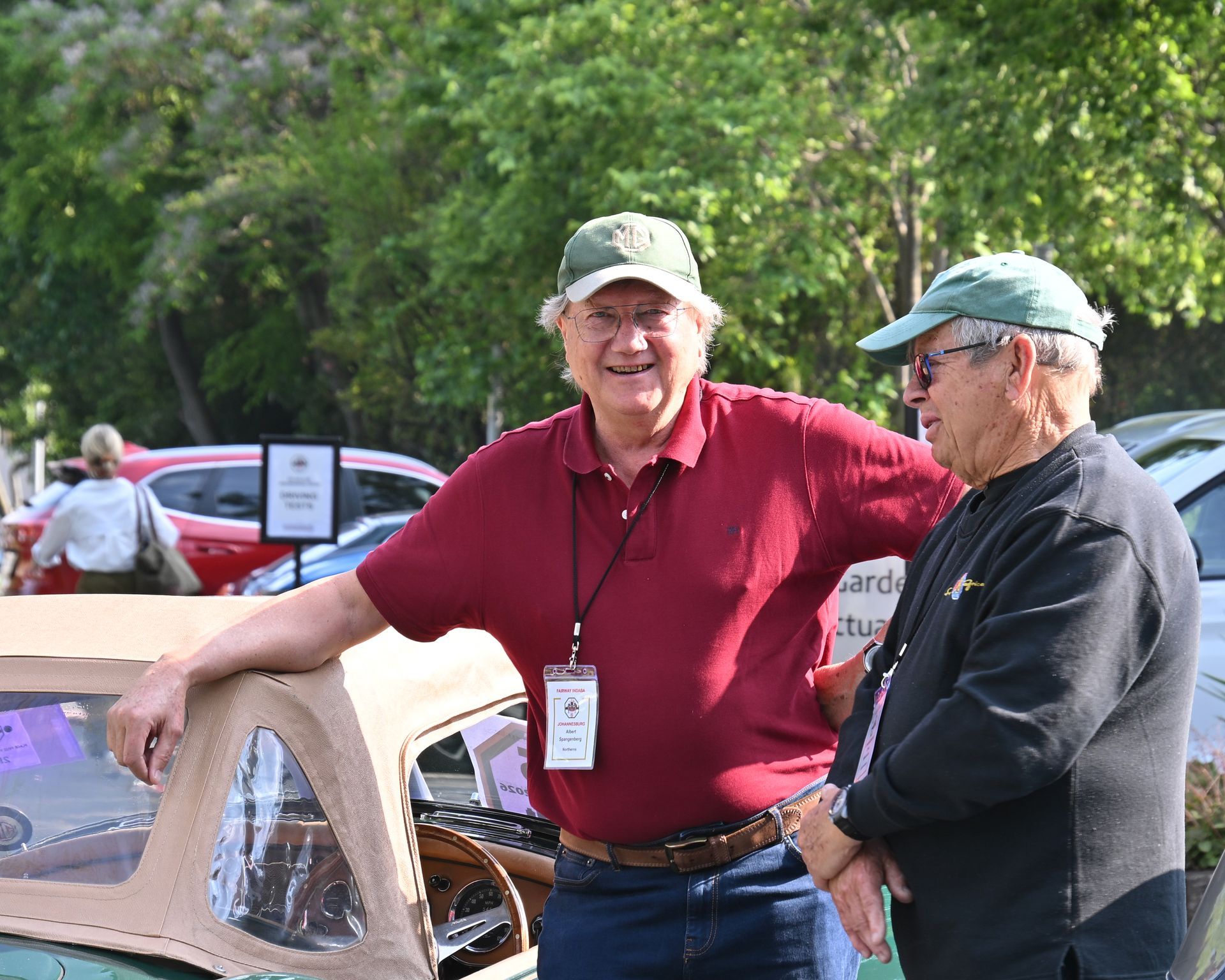 Two men in caps chatting outdoors beside a vintage car, one leaning on the car with a red shirt.