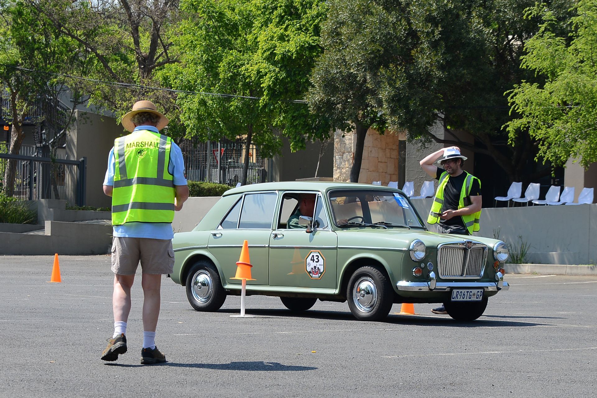 Vintage green car driving through traffic cones with two safety marshals nearby outdoors