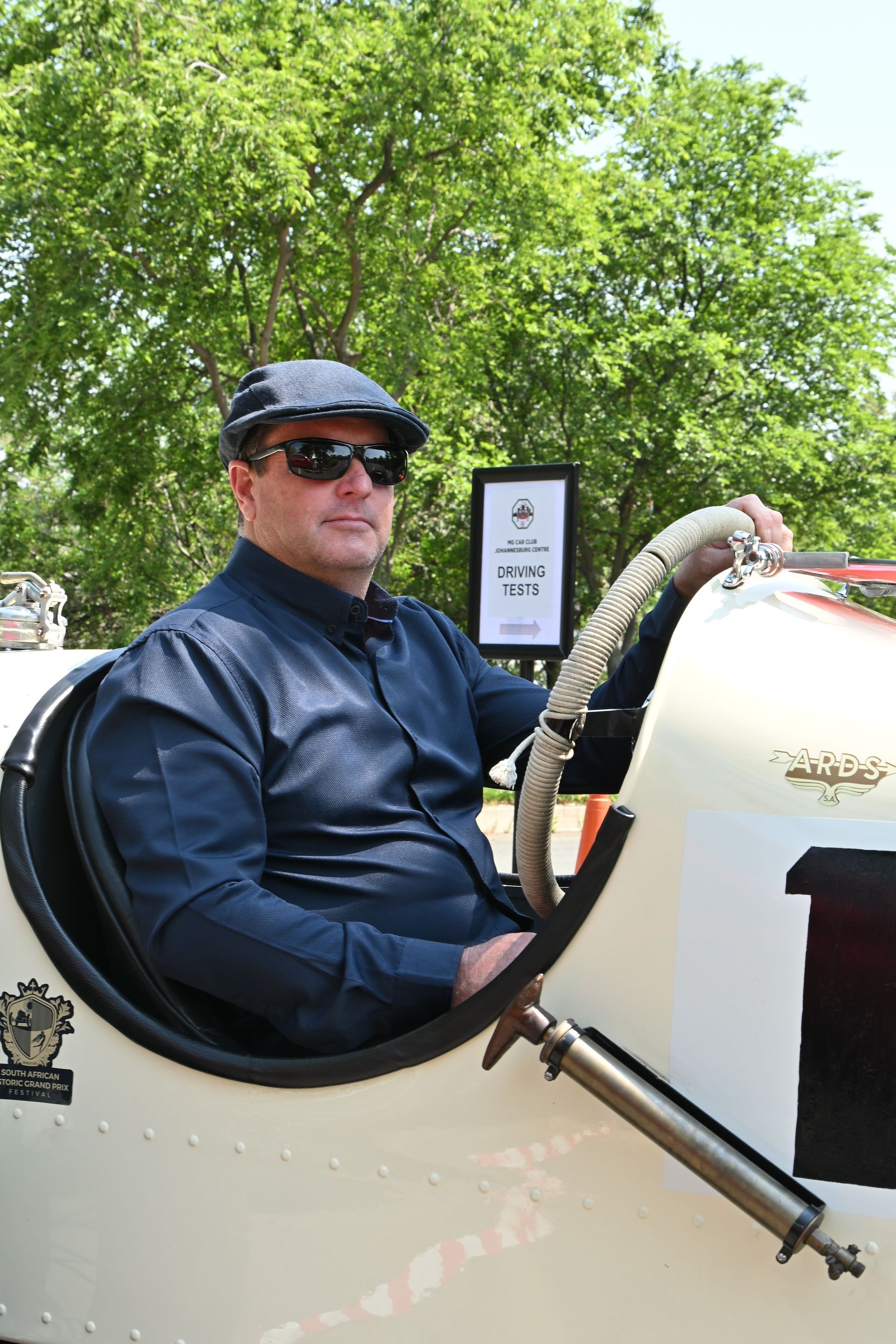 Man in dark jacket and cap sitting in a white open vehicle outdoors, with green trees behind him