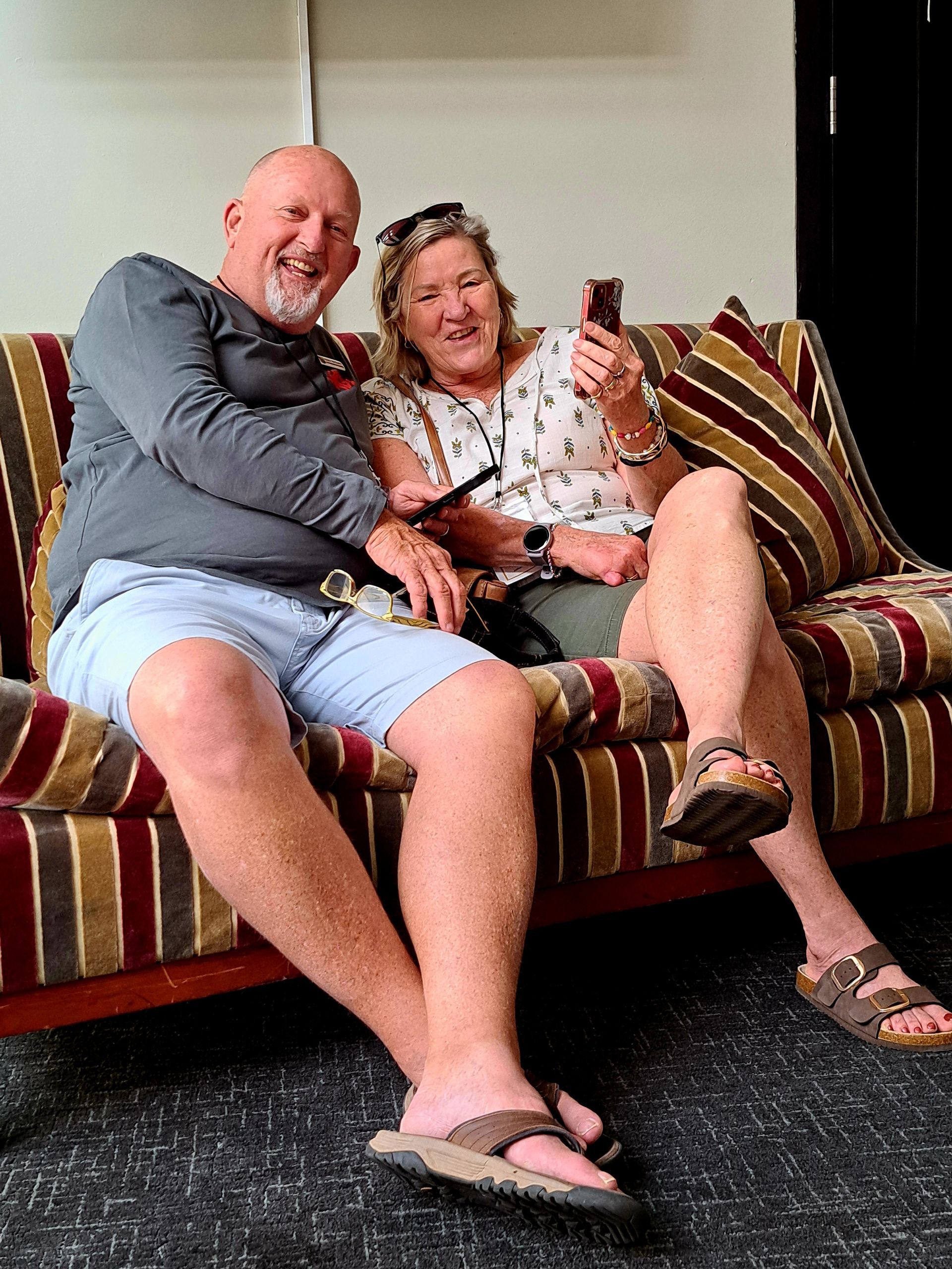 Two people smiling on a striped couch, one holding a phone, in a cozy living room.