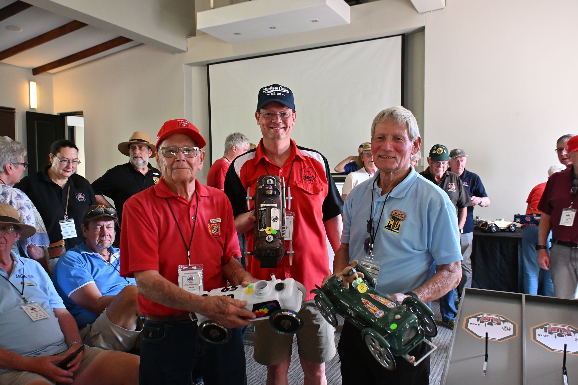 Three men pose indoors with model trucks, one holding a trophy, at a hobby or awards event.