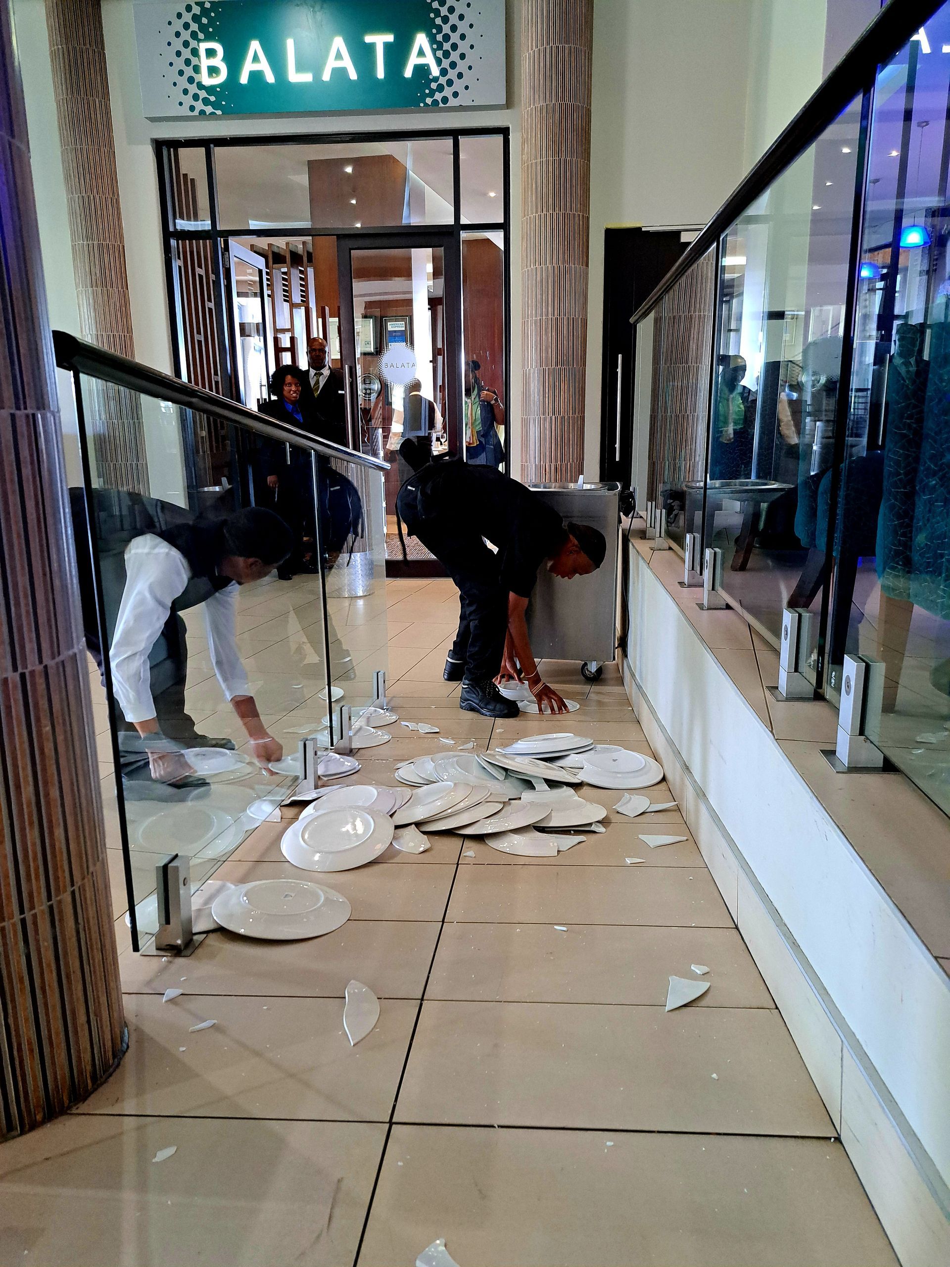 Two people cleaning broken plates in a mall corridor outside a store named BALATA