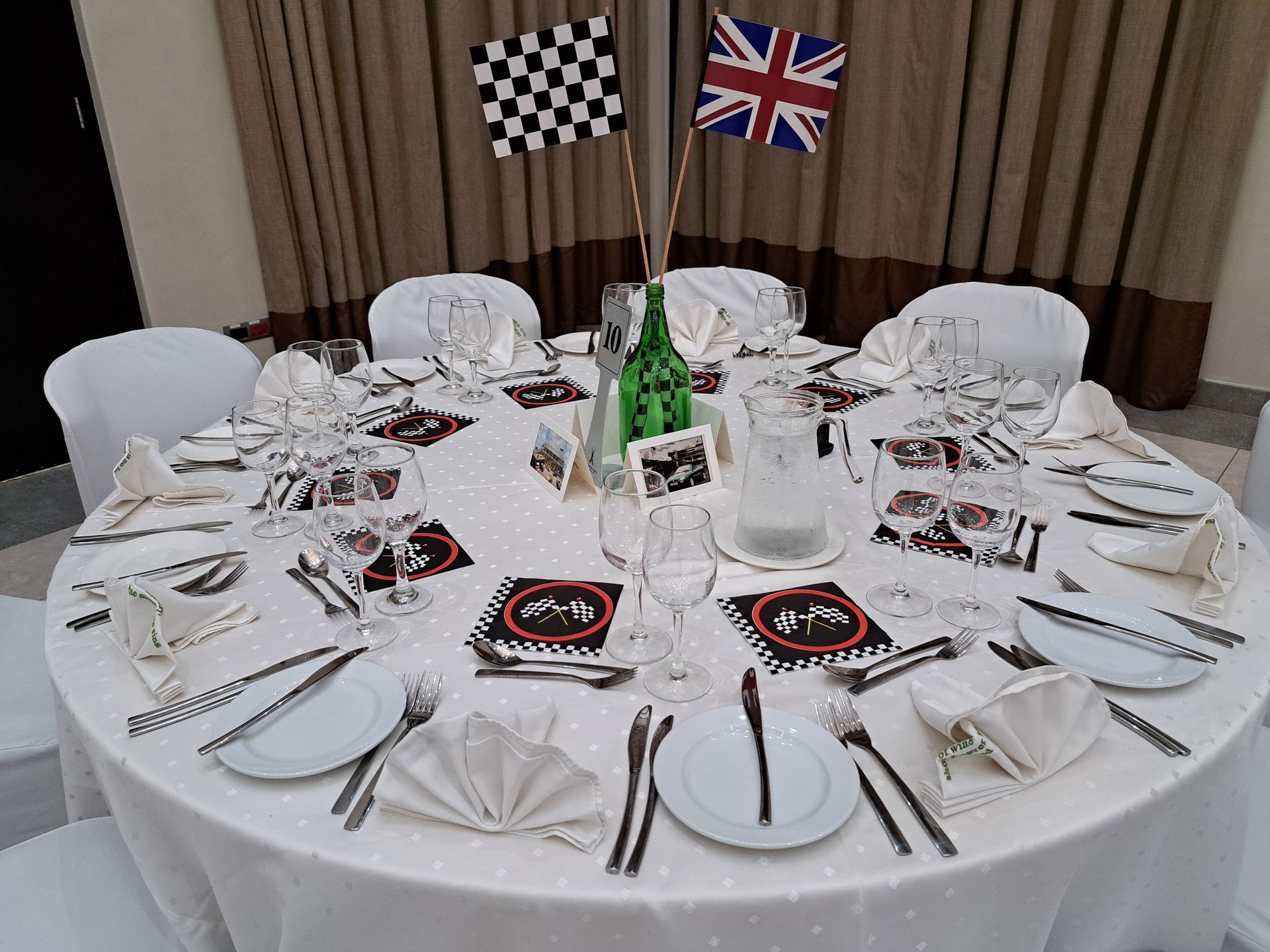 Round banquet table set with white plates, glassware, center bottle, and UK and checkered flags in the background