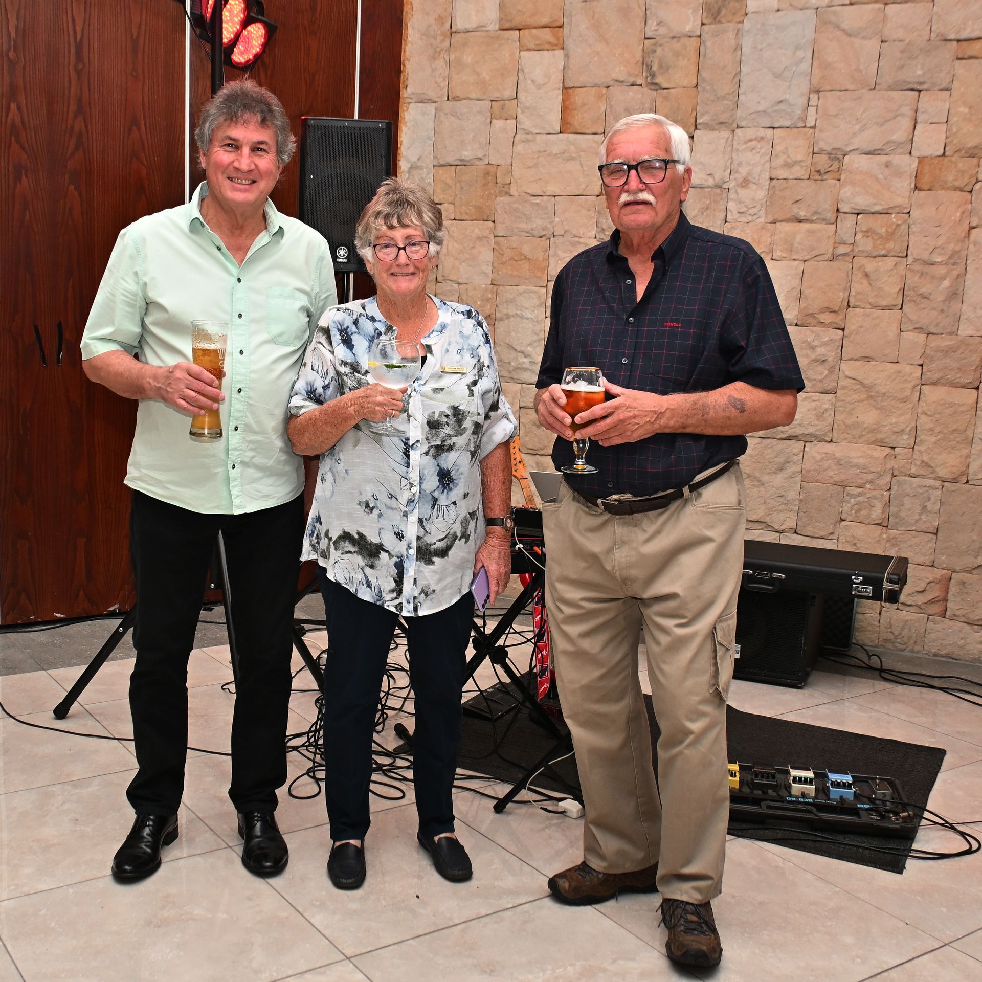 Three people standing with drinks in front of a stone wall, posing at an indoor event.