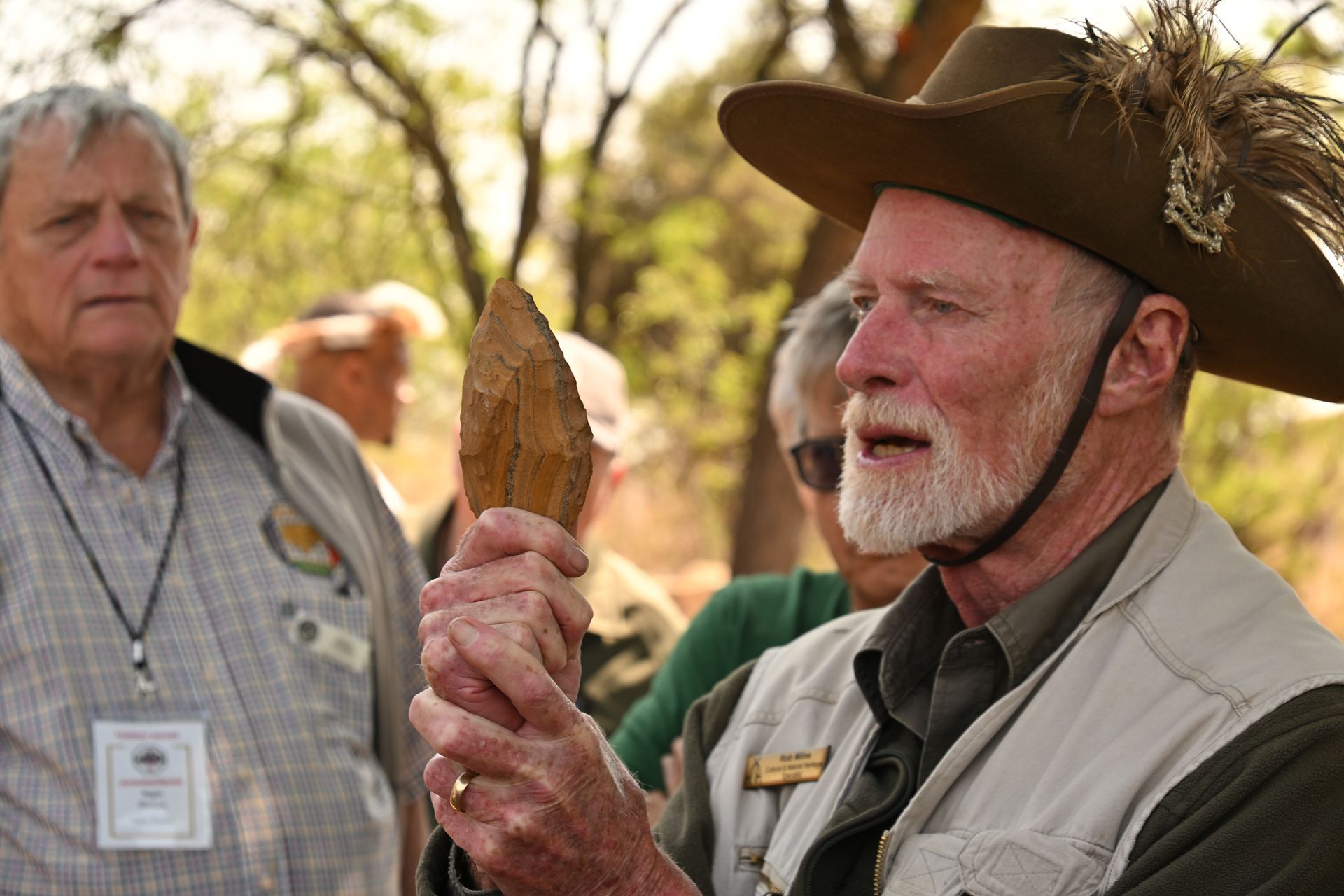 Man in brown hat holding a stone artifact outdoors, with another man in the background