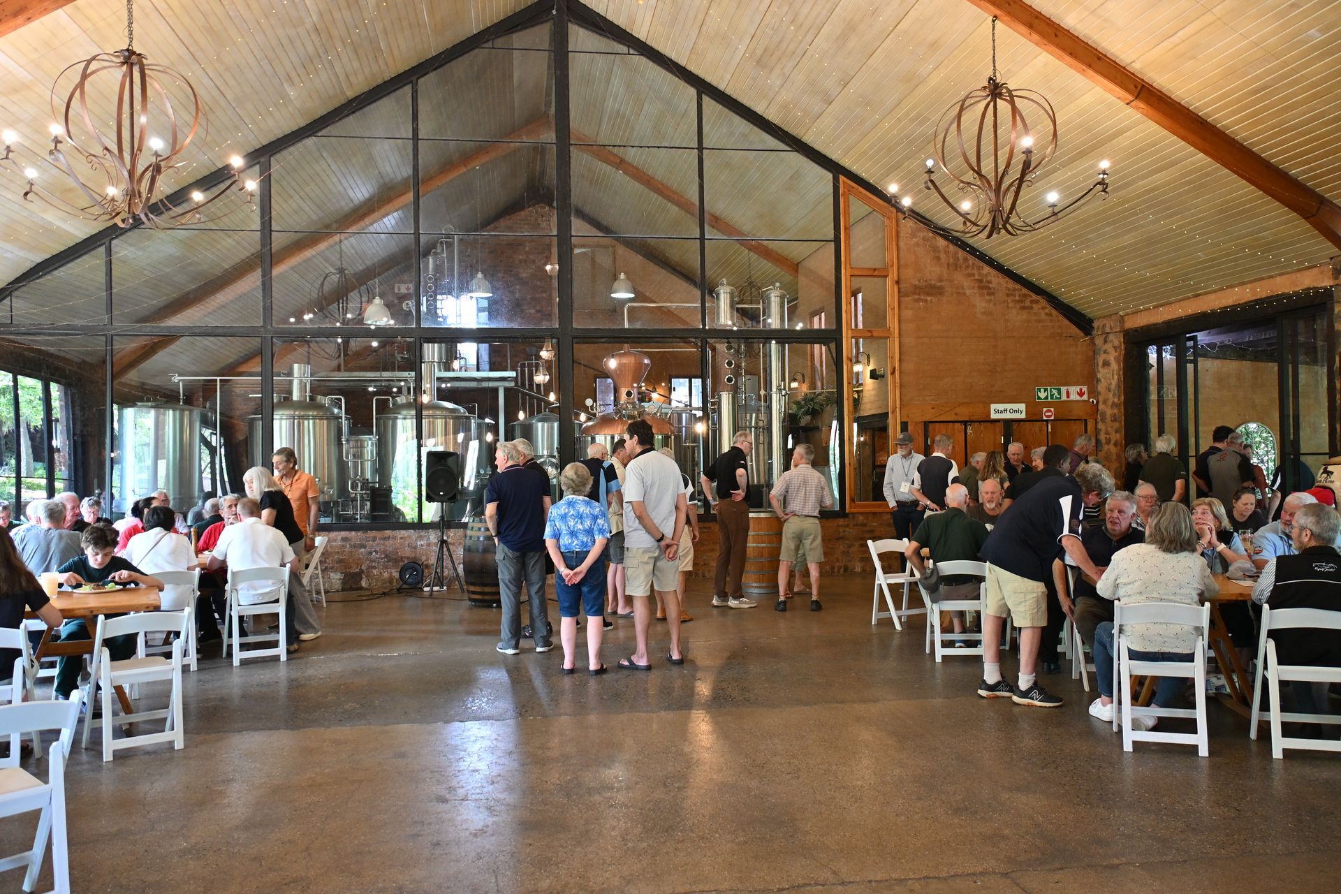 Crowded rustic banquet hall with guests seated at tables and others standing near a glass-walled entrance