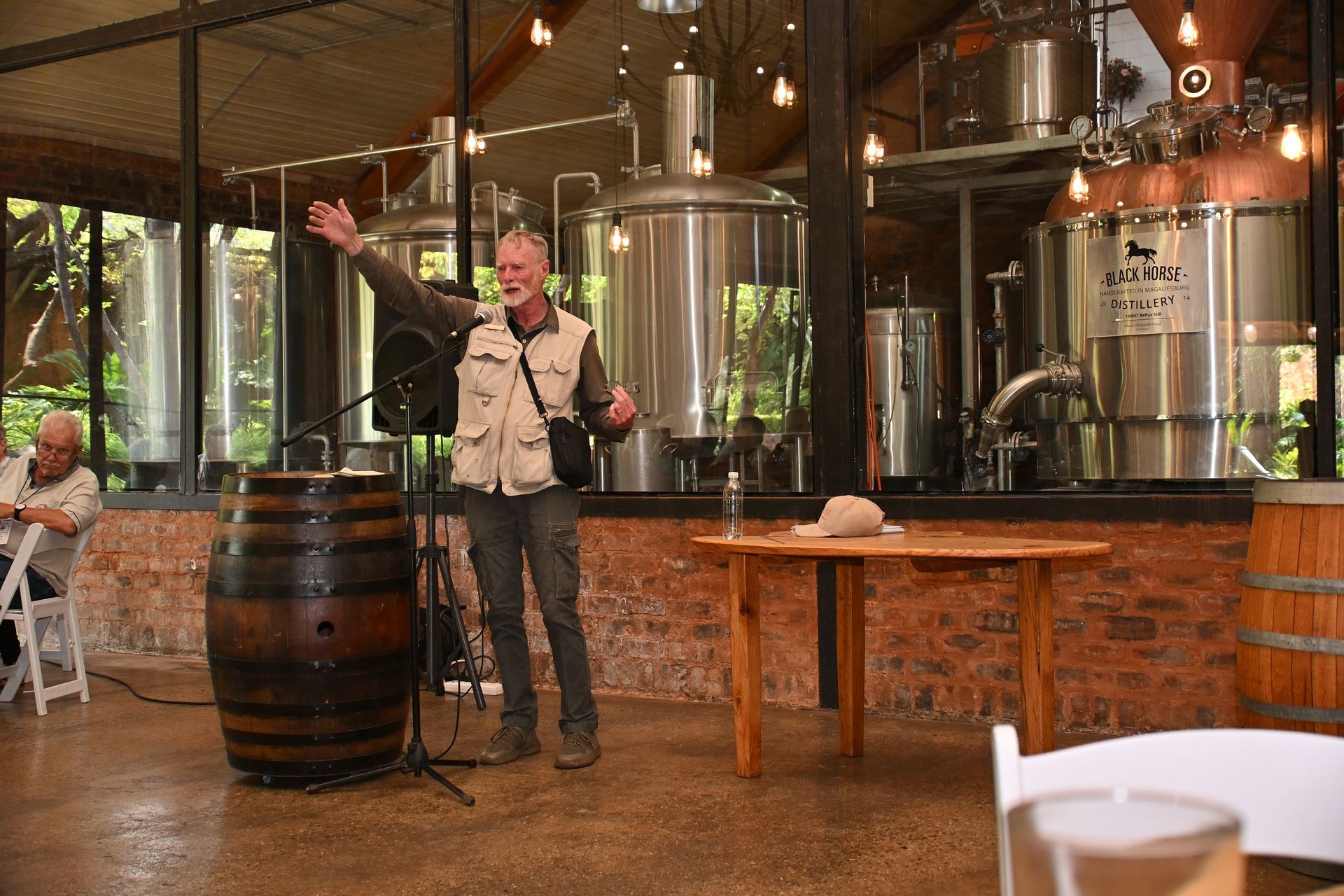 Person speaking inside a brewery, standing by stainless steel tanks and wooden barrels.