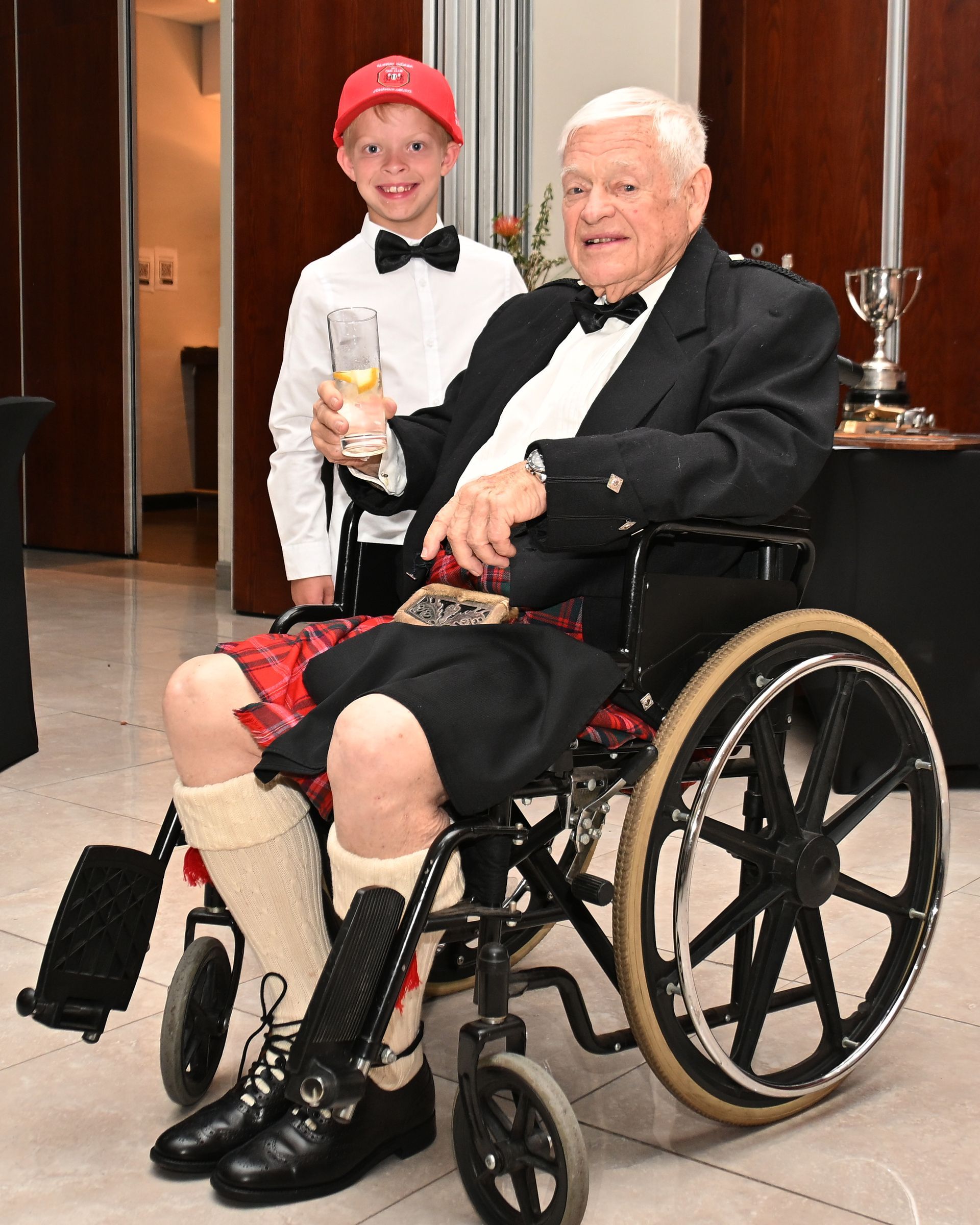 Man in a wheelchair holding a trophy, with a smiling person standing behind him in a formal indoor setting.