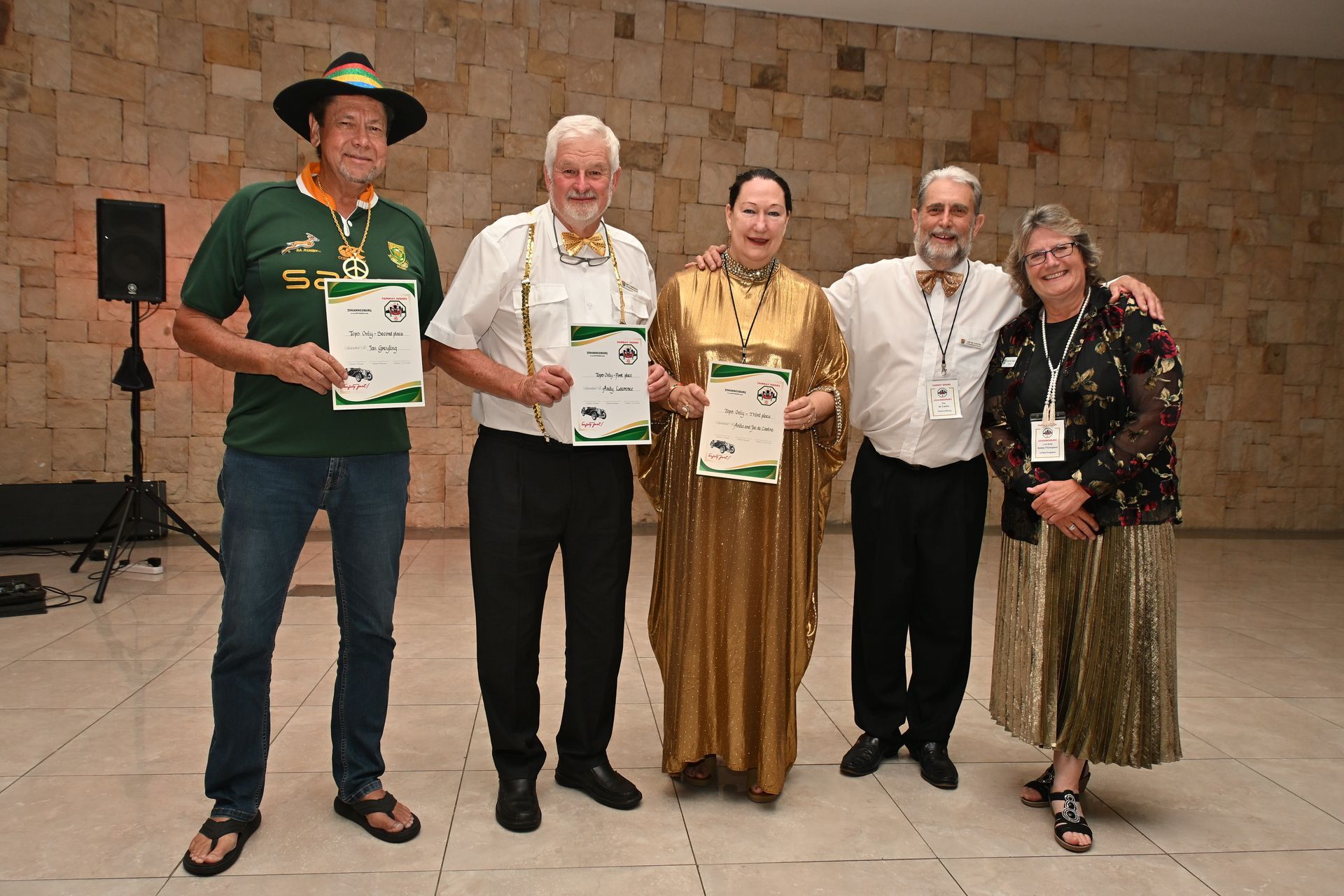 Five people stand holding certificates in a stone-walled room, posing for a group photo.
