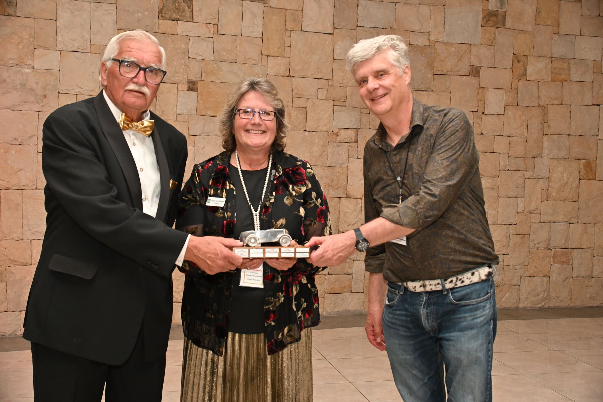 Three people holding a plaque at an award presentation in front of a brick wall.