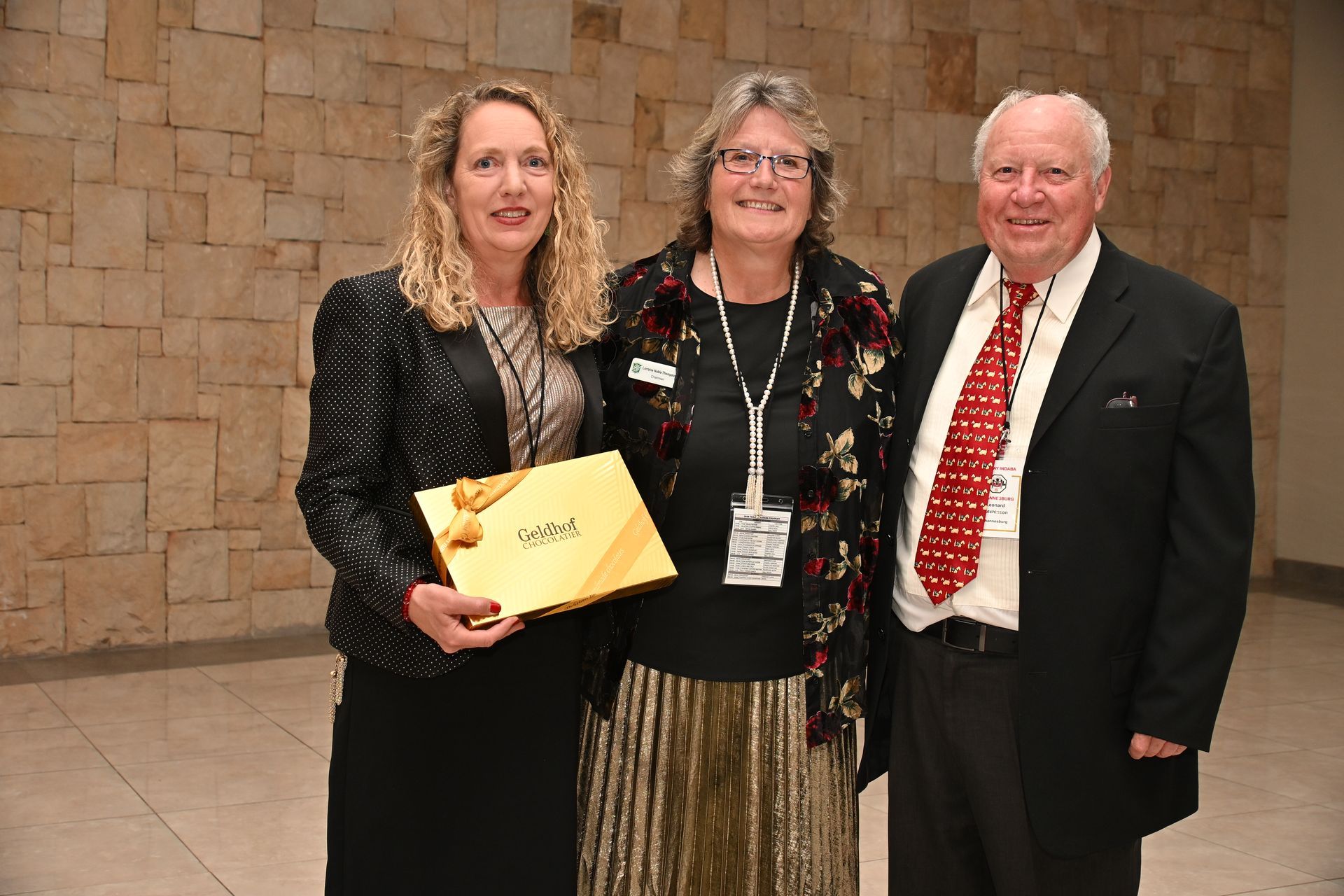 Three people posing indoors, holding a yellow folder, in front of a stone wall.