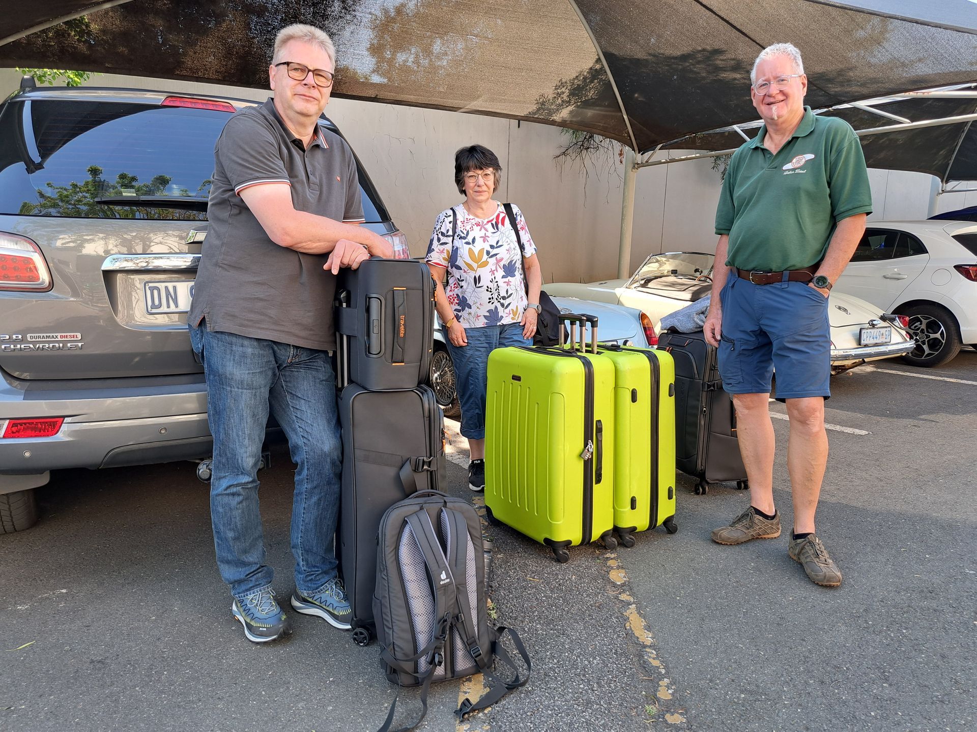 Three people standing with luggage and backpacks in a parking lot under a covered area.