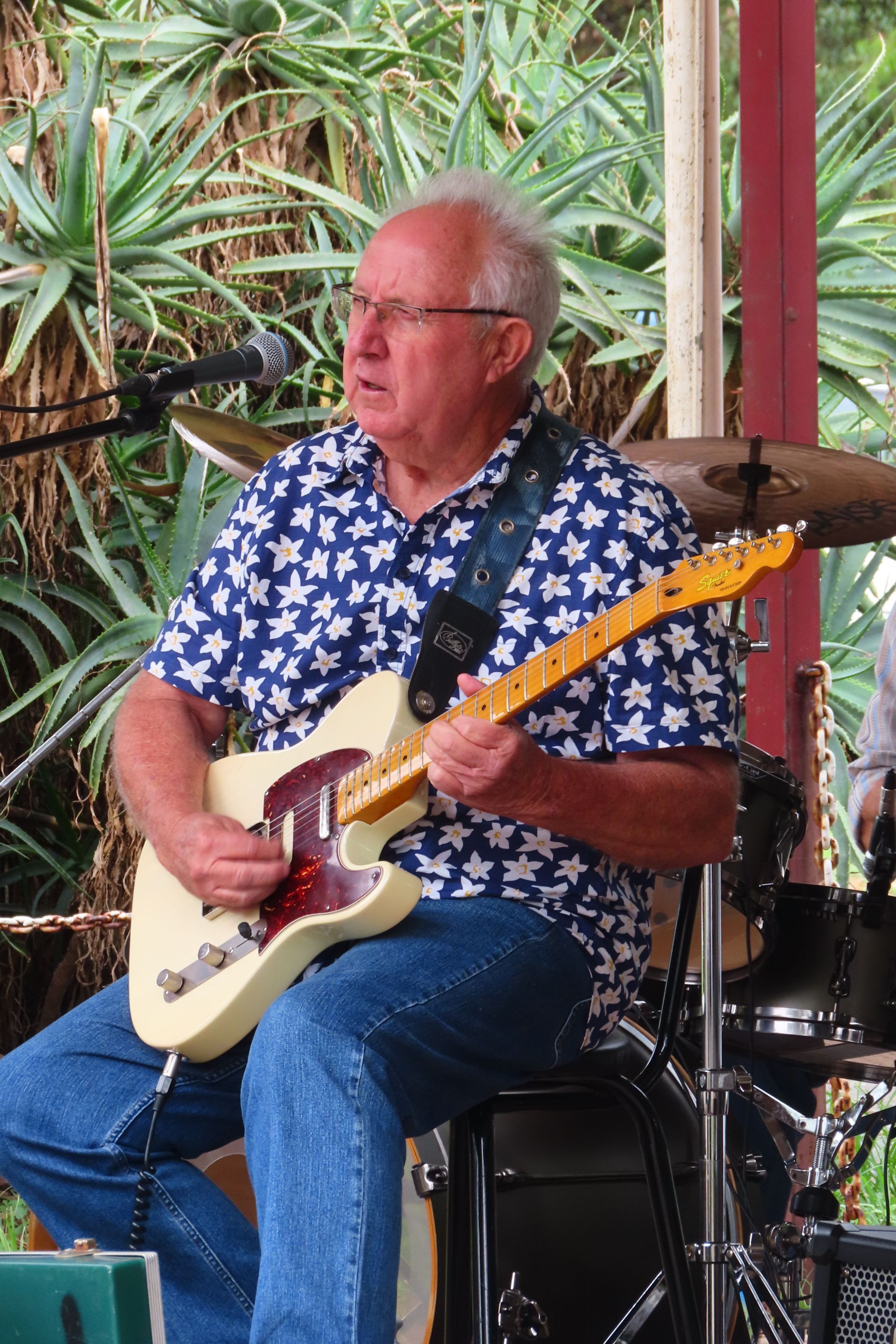 Man playing a white electric guitar, sitting, wearing blue star-patterned shirt and jeans, outdoor setting.