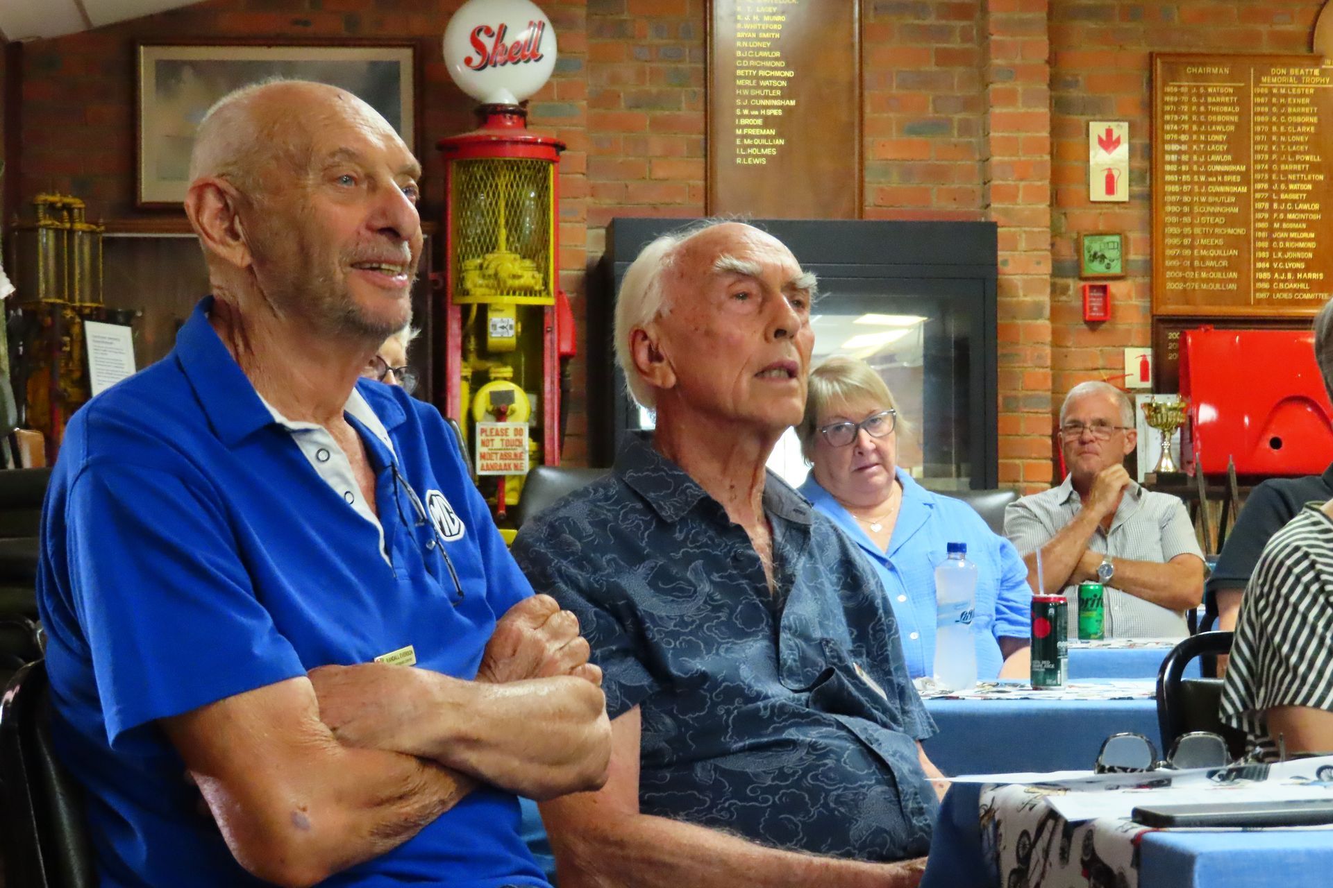 Two older men seated at a table, looking to their right. Brick wall with vintage gas pump in background.