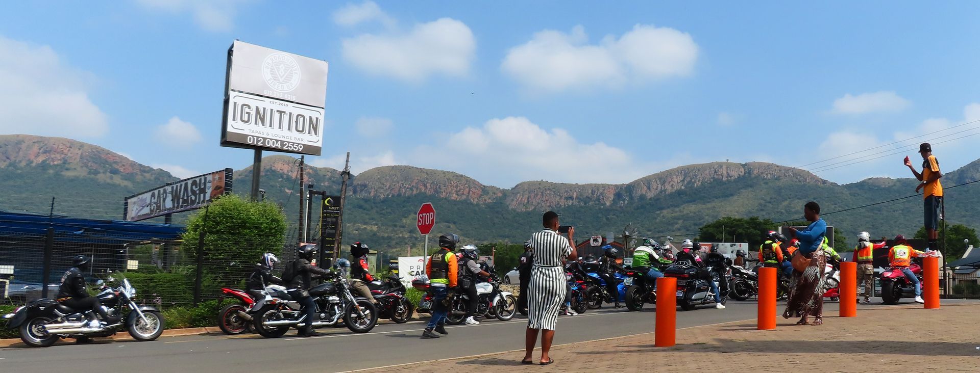 Motorcycles parked in front of a sign, people standing around. Mountains in background, blue sky.