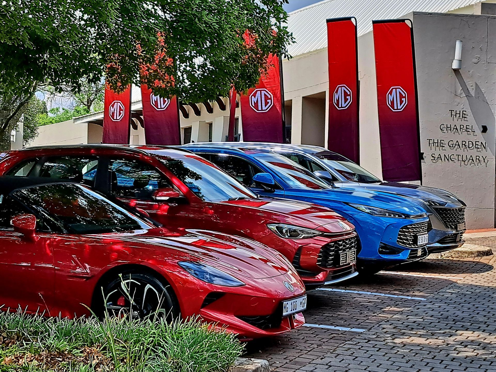 Row of red and blue sports cars parked outside a building with red banners and trees
