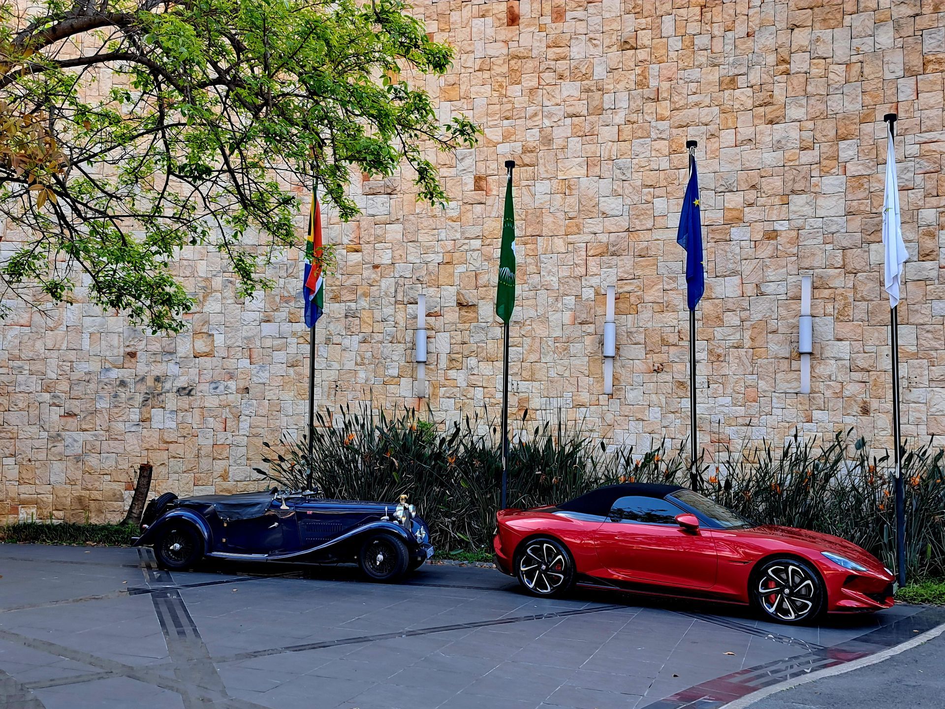 Two sports cars, one black and one red, parked beside a stone wall with flags overhead