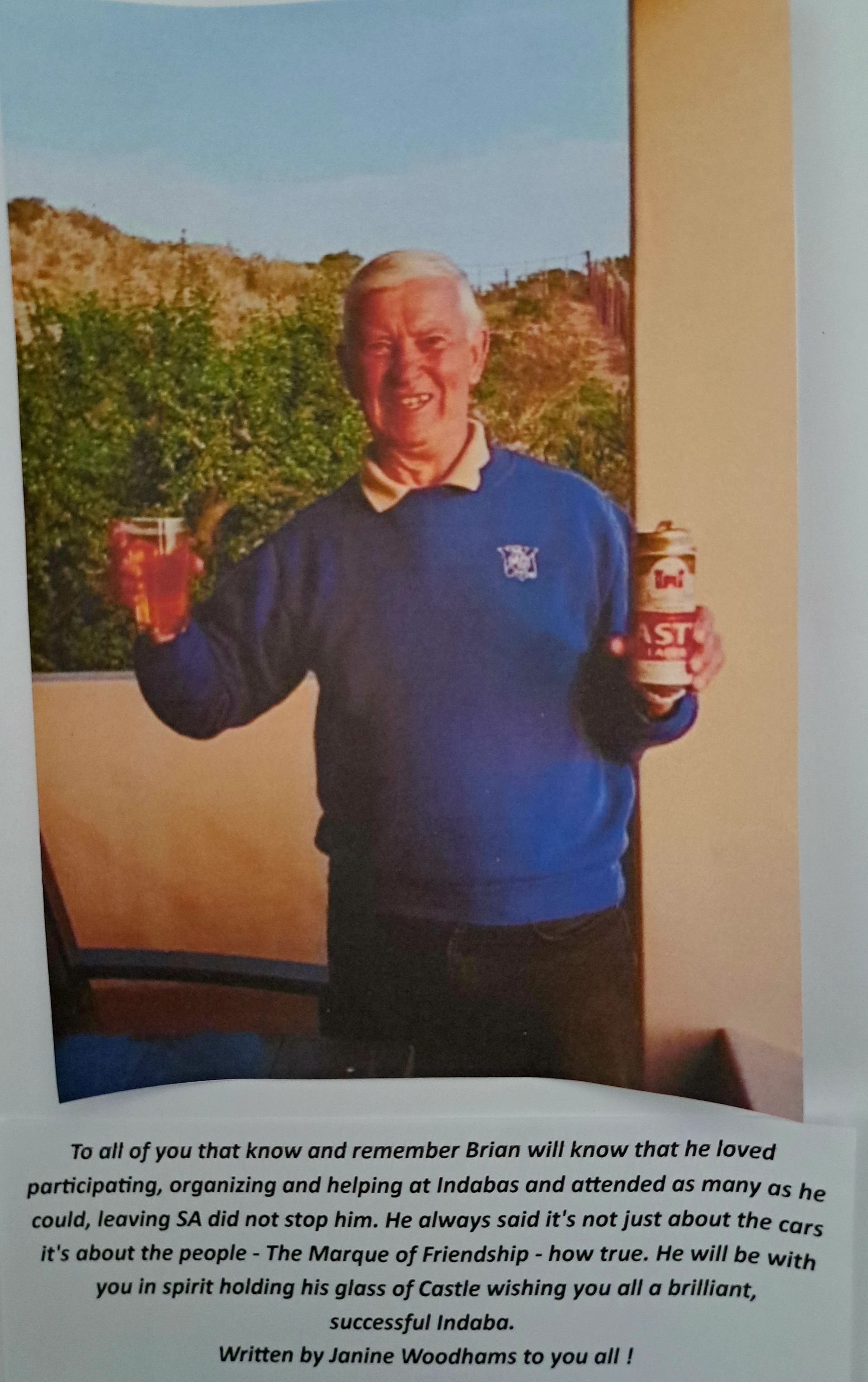 Man in blue sweater holding two cups, standing indoors beside a wall with a landscape photo behind him.