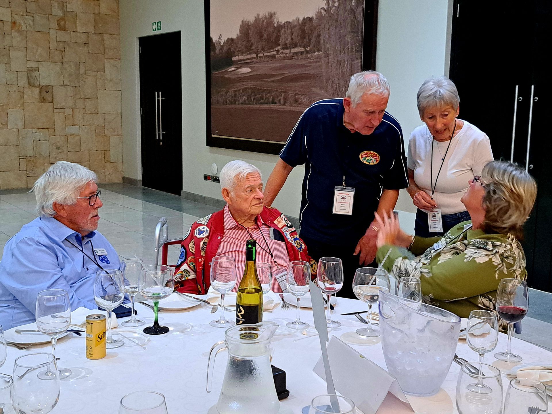 Four people chatting around a banquet table in a bright dining hall, with wine and folded napkins.