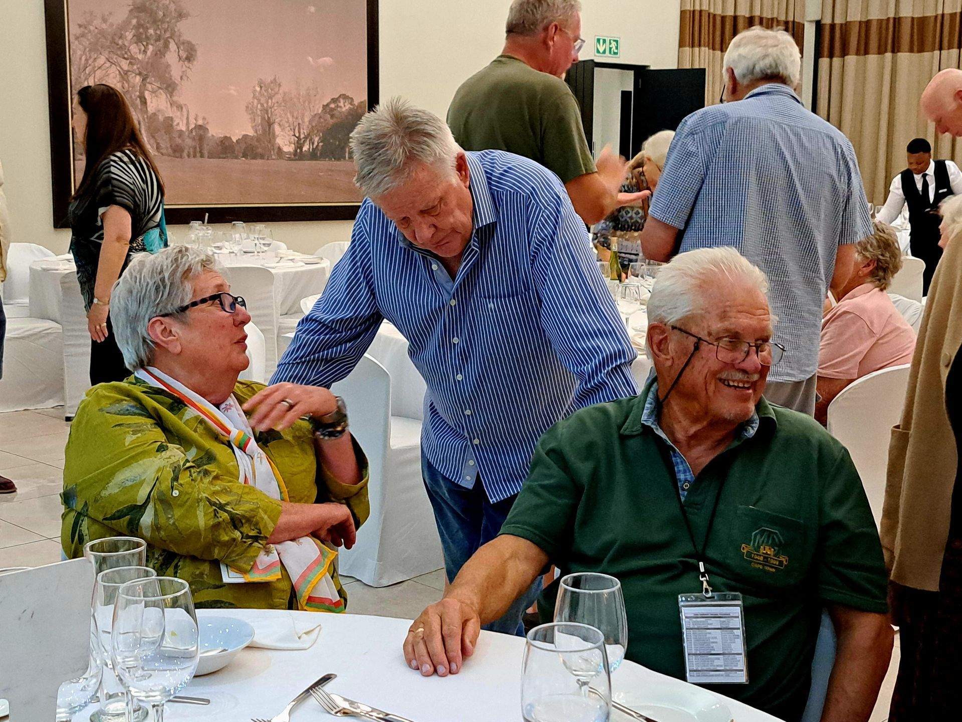 Three older adults chatting at a banquet table in a bright dining hall