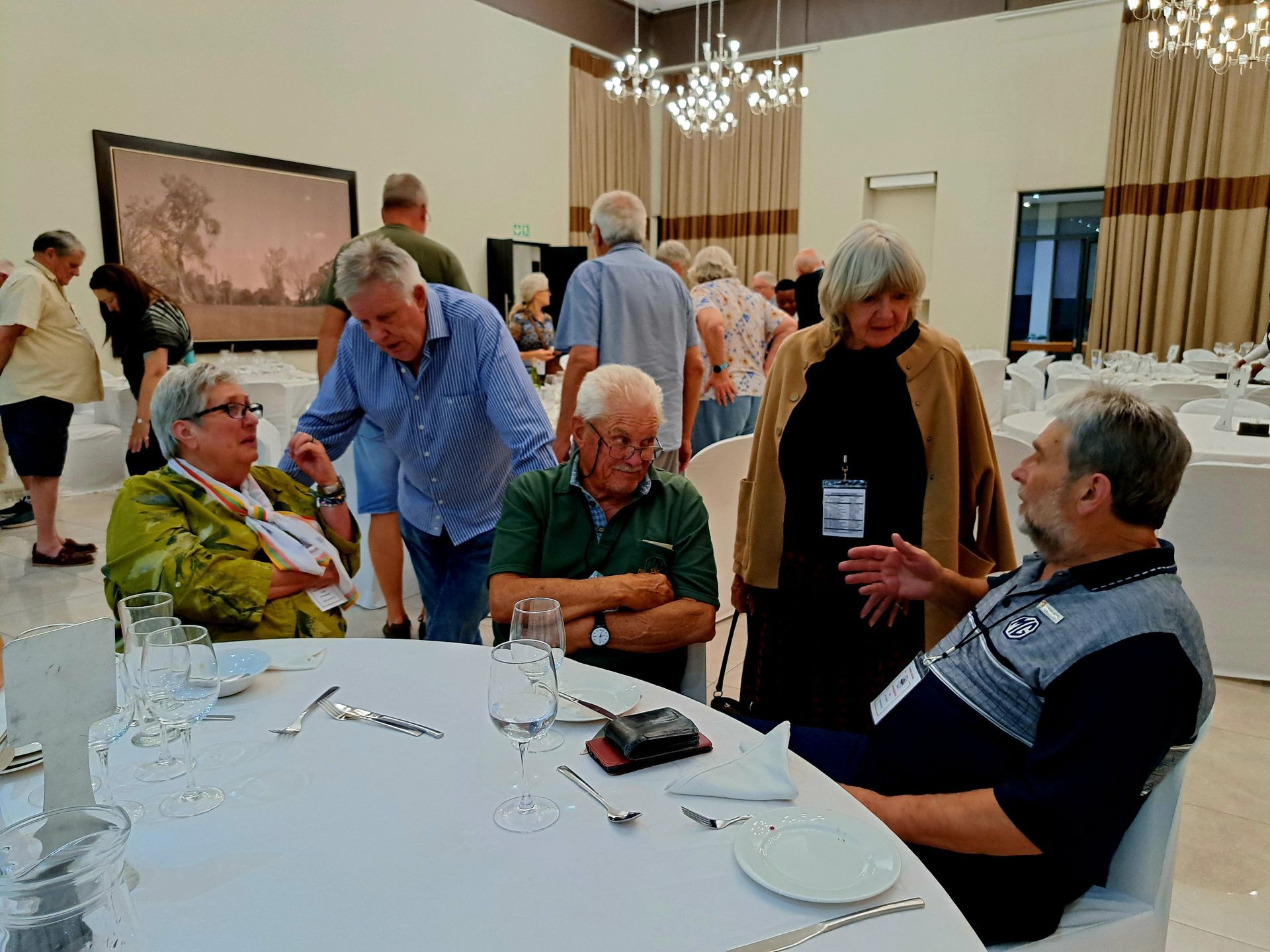 People mingling around a banquet table in a bright dining room with chandeliers.