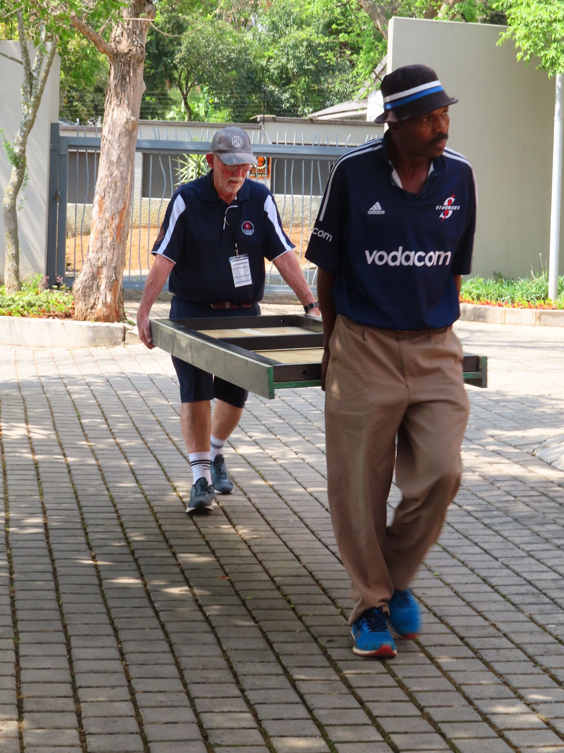 Two workers carry a large tray along a paved outdoor walkway near a building entrance.