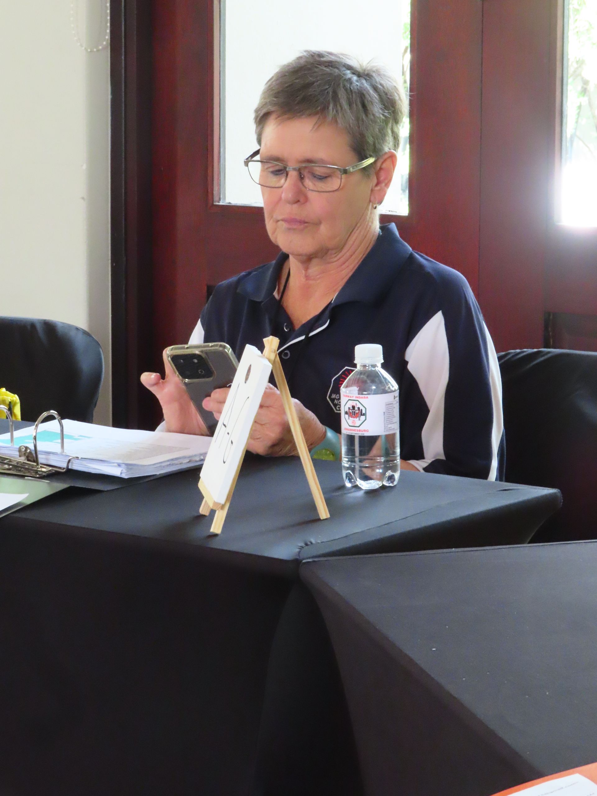 Woman reading a phone at a table with papers, a water bottle, and a nameplate in a meeting room