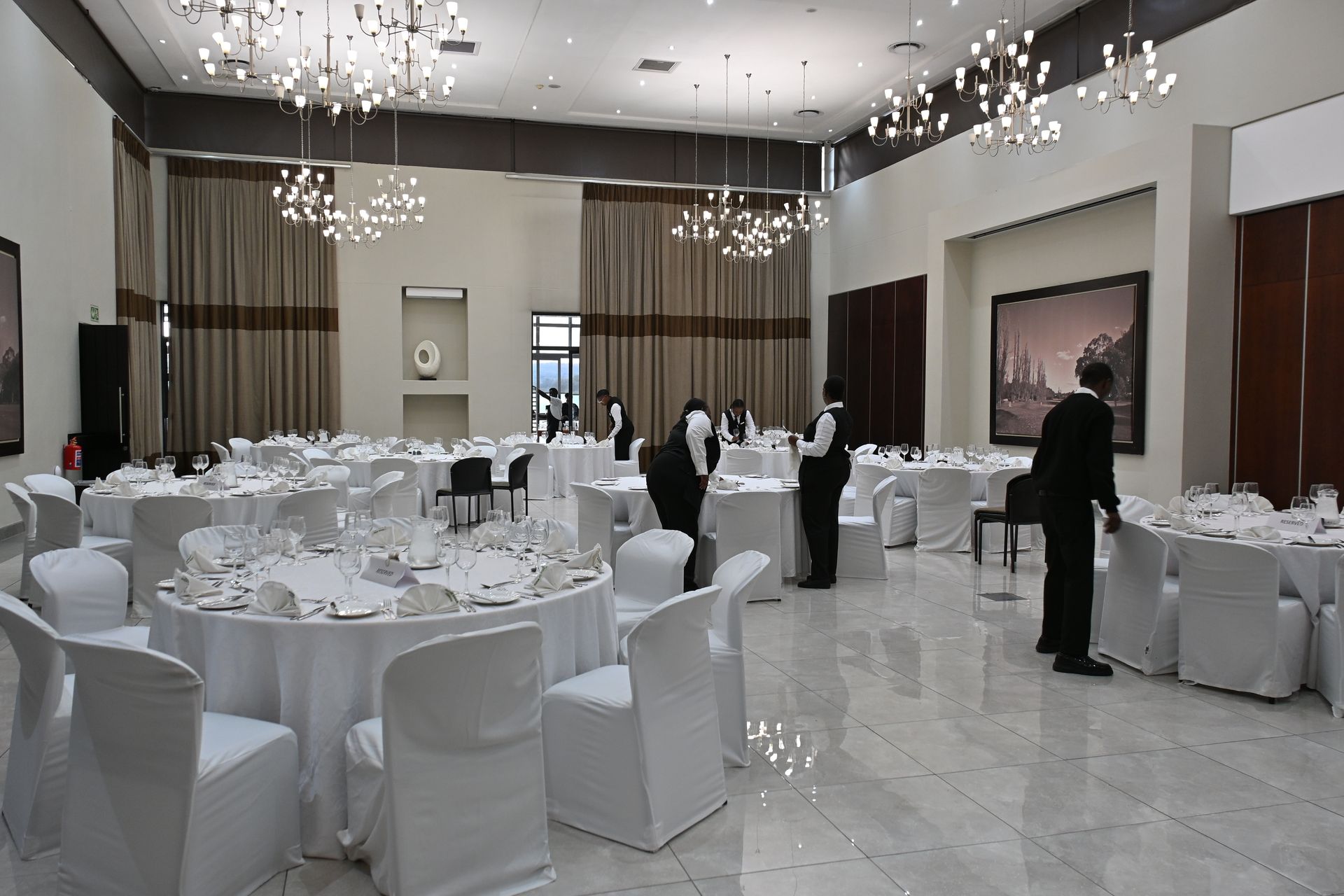 Elegant banquet hall set with round tables, white chair covers, chandeliers, and a few guests standing near the back