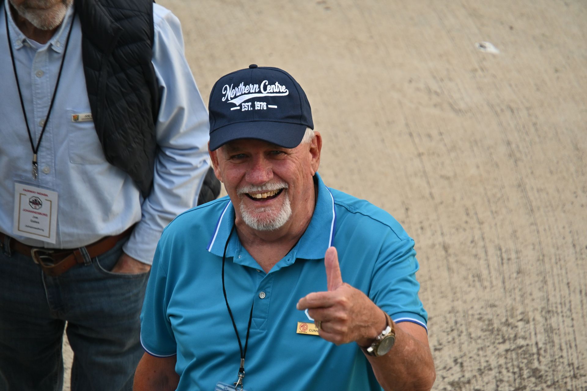 Smiling man in a blue polo and cap gives a thumbs-up on a dirt track