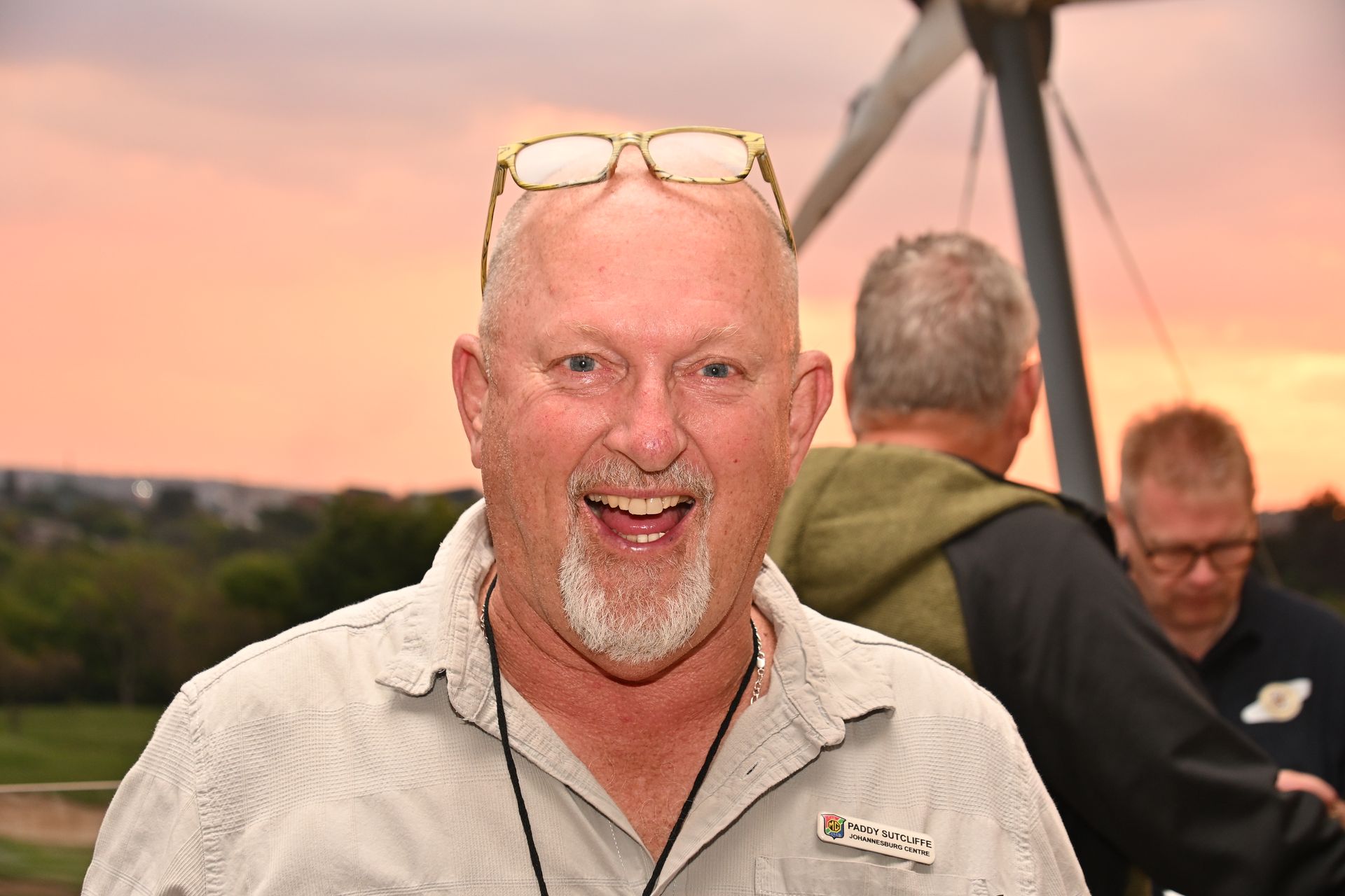 Smiling man in beige shirt with glasses on his head at sunset, with others and a bridge in the background