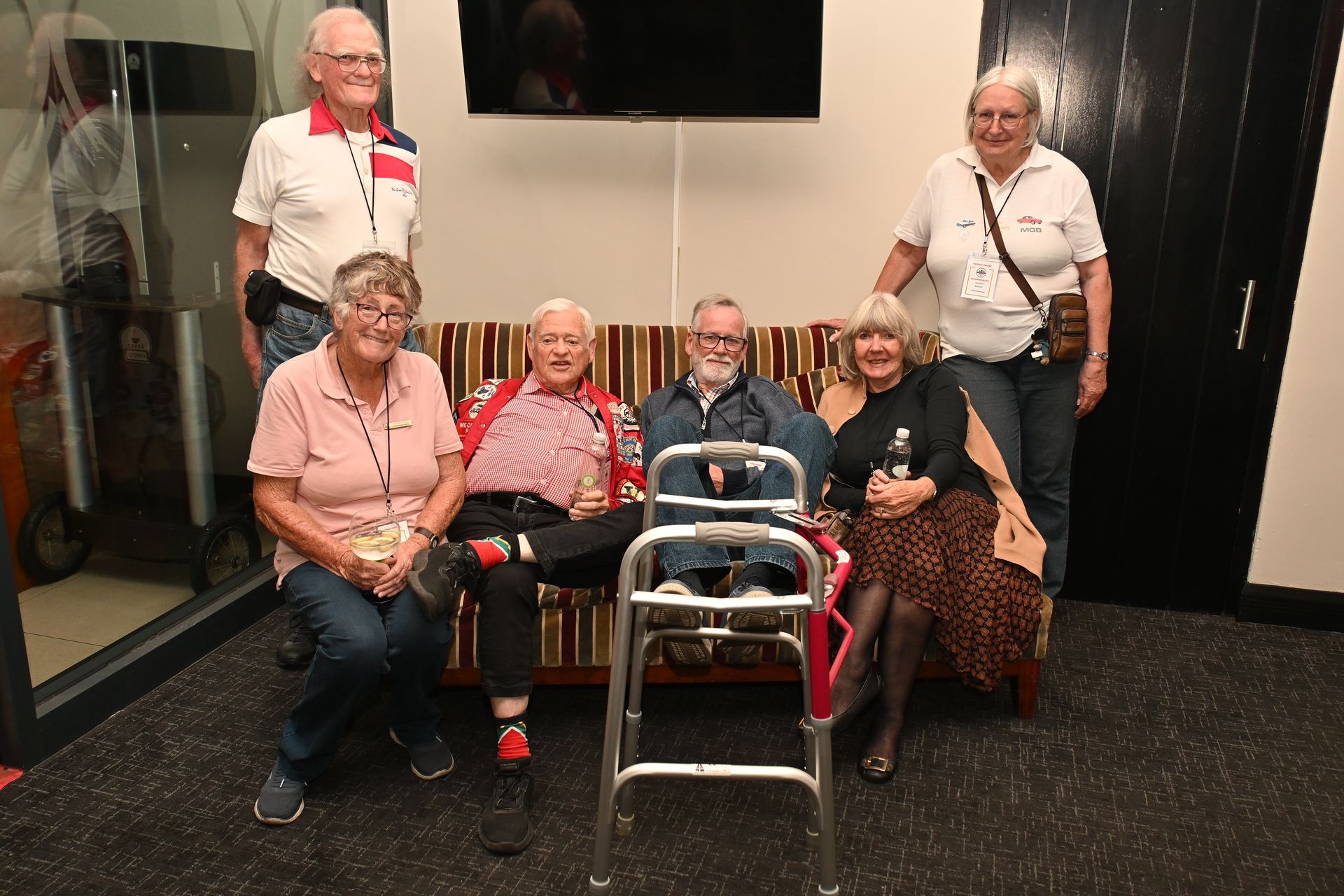 Six people posing indoors, with one standing and five seated around a walker.