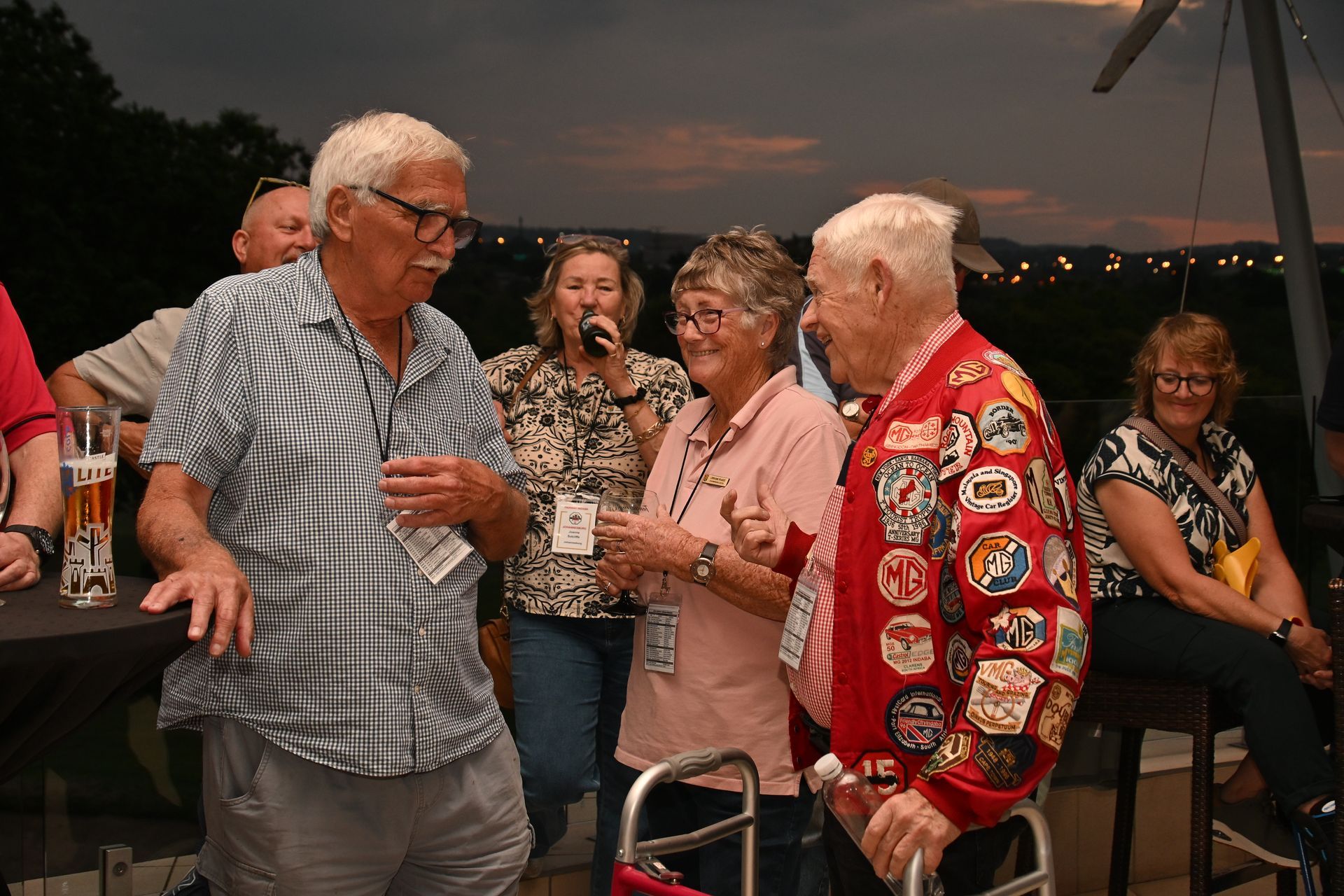 People socializing on a patio at dusk, holding drinks and smiling near a waterfront view