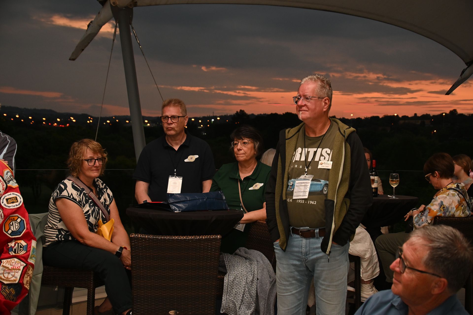 People standing outdoors at sunset, gathered around a table on a deck with city lights in the background