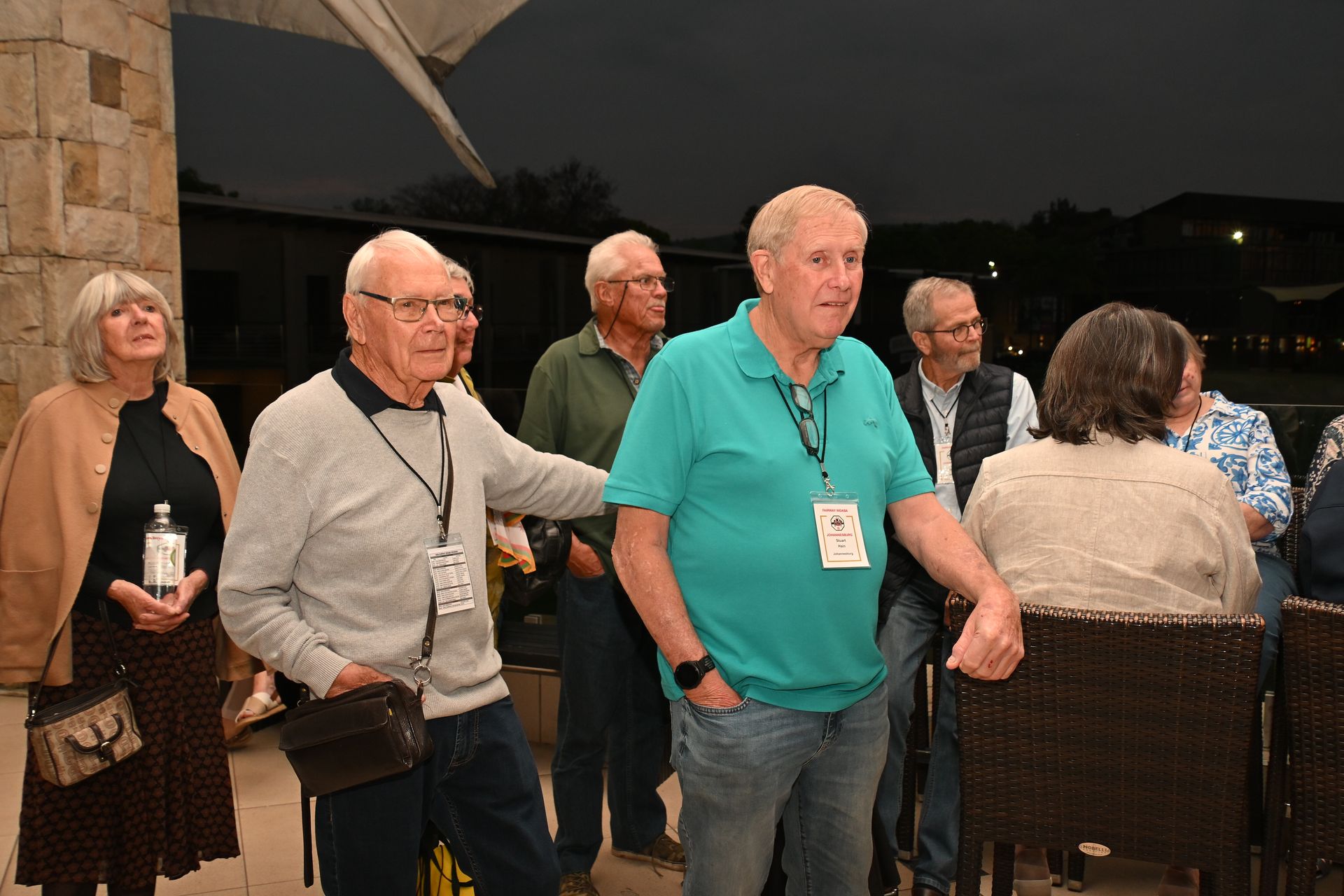 People standing and chatting at an outdoor evening gathering, with a man in a teal shirt in the foreground.