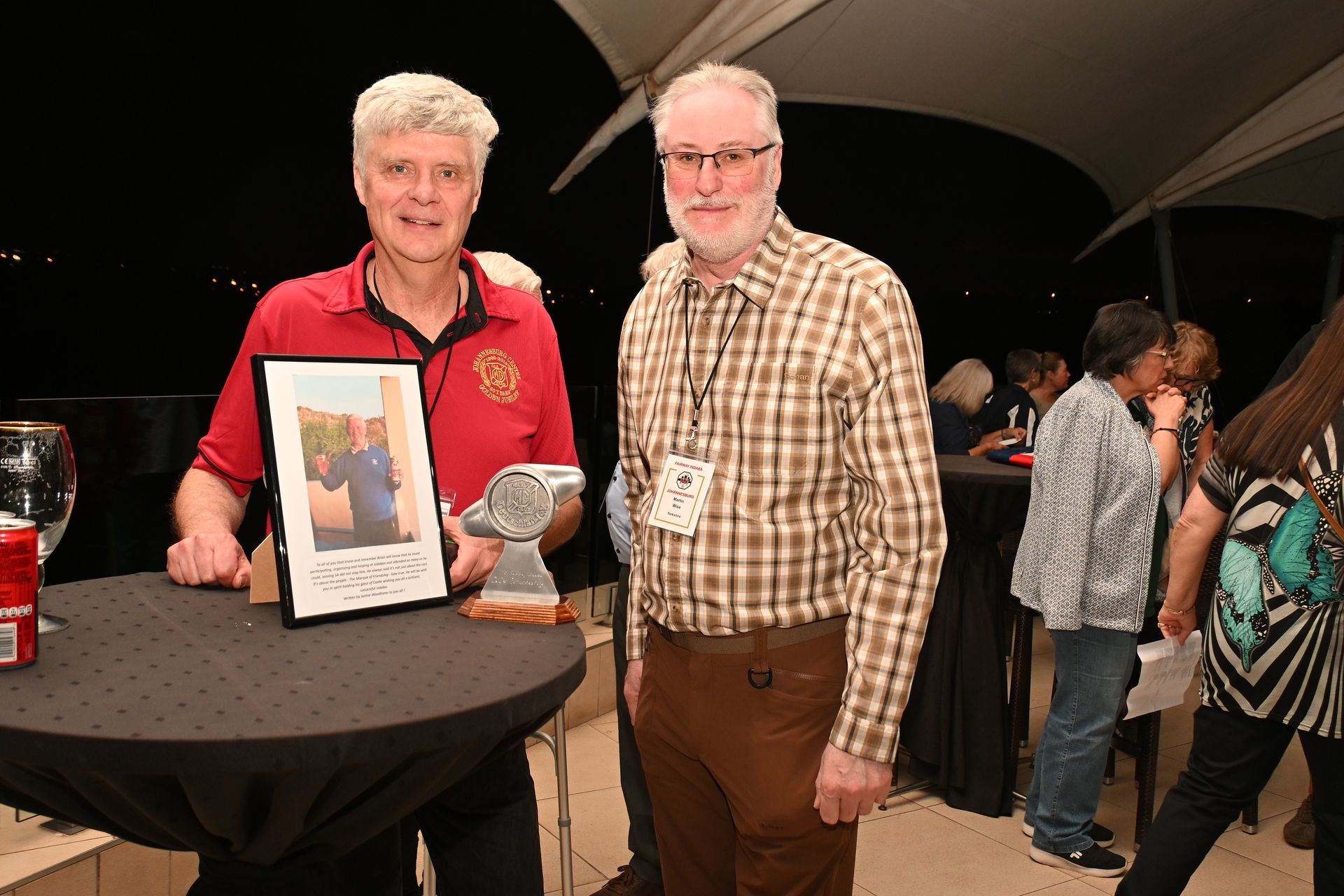 Two men standing at a table with a framed photo and plaque at an evening event under a tent