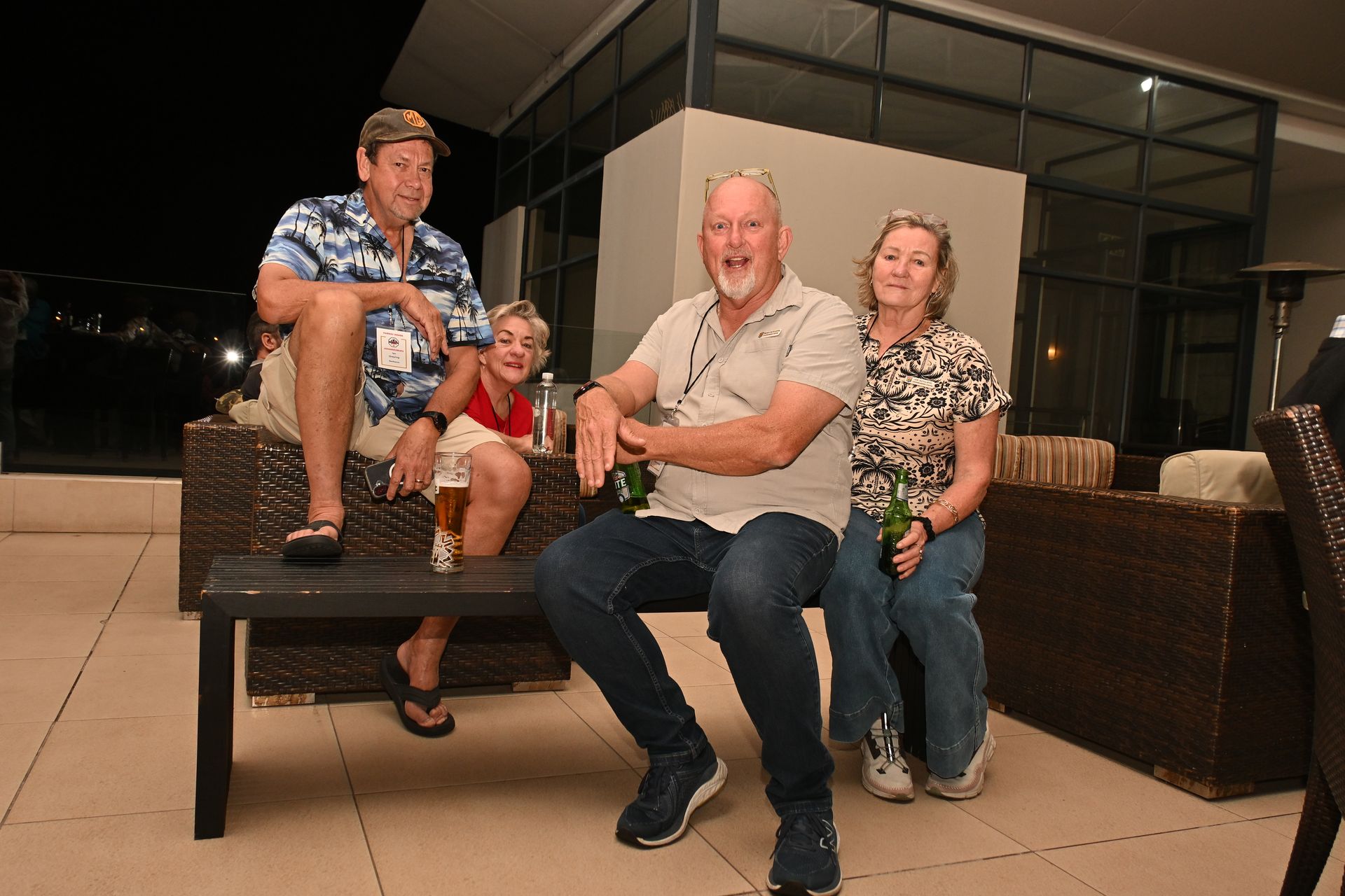 Four people sit on a wicker patio bench at night, smiling in a casual outdoor setting.