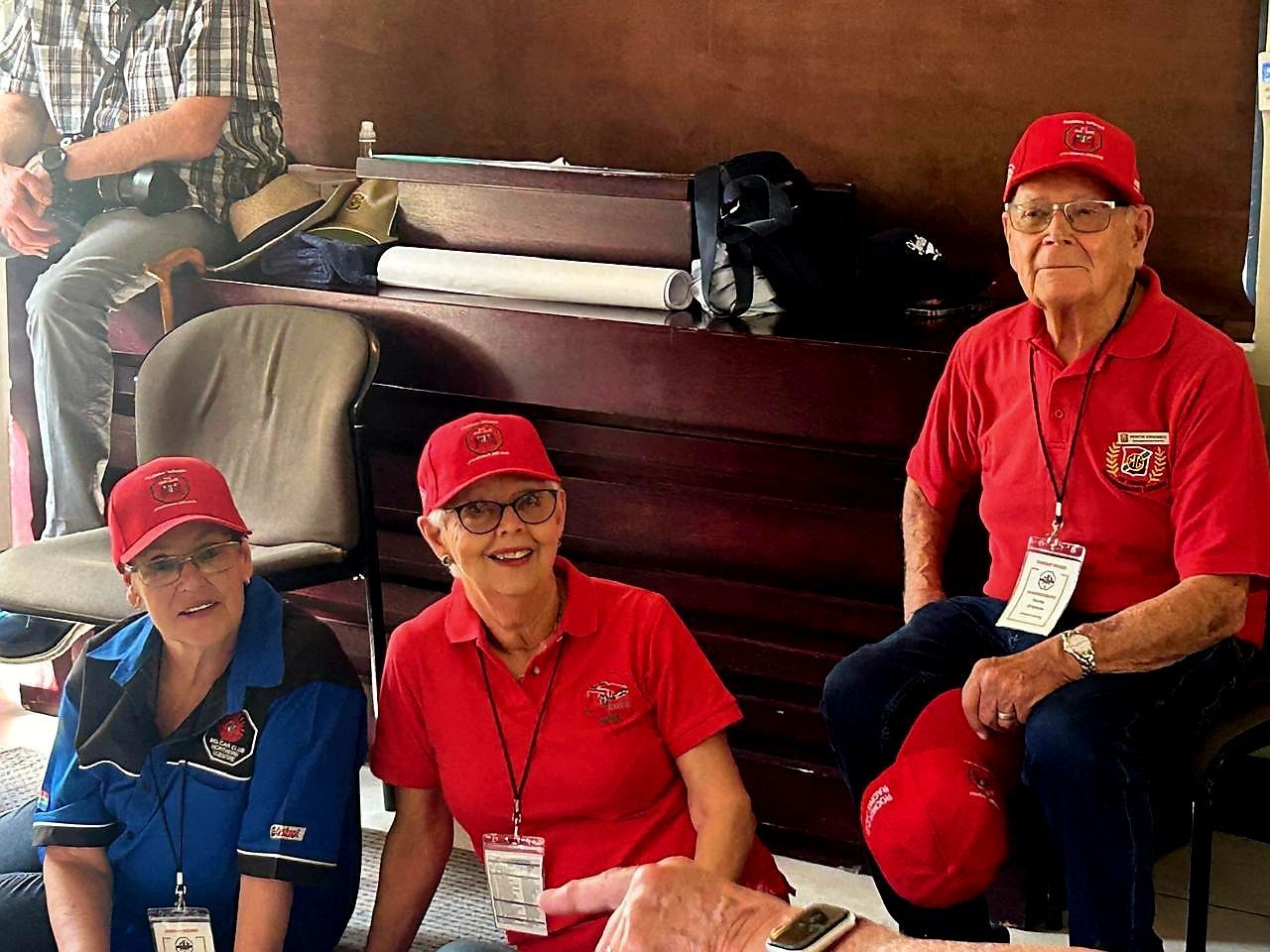 Three people in red caps and shirts sit indoors, smiling near a stack of folded blankets.
