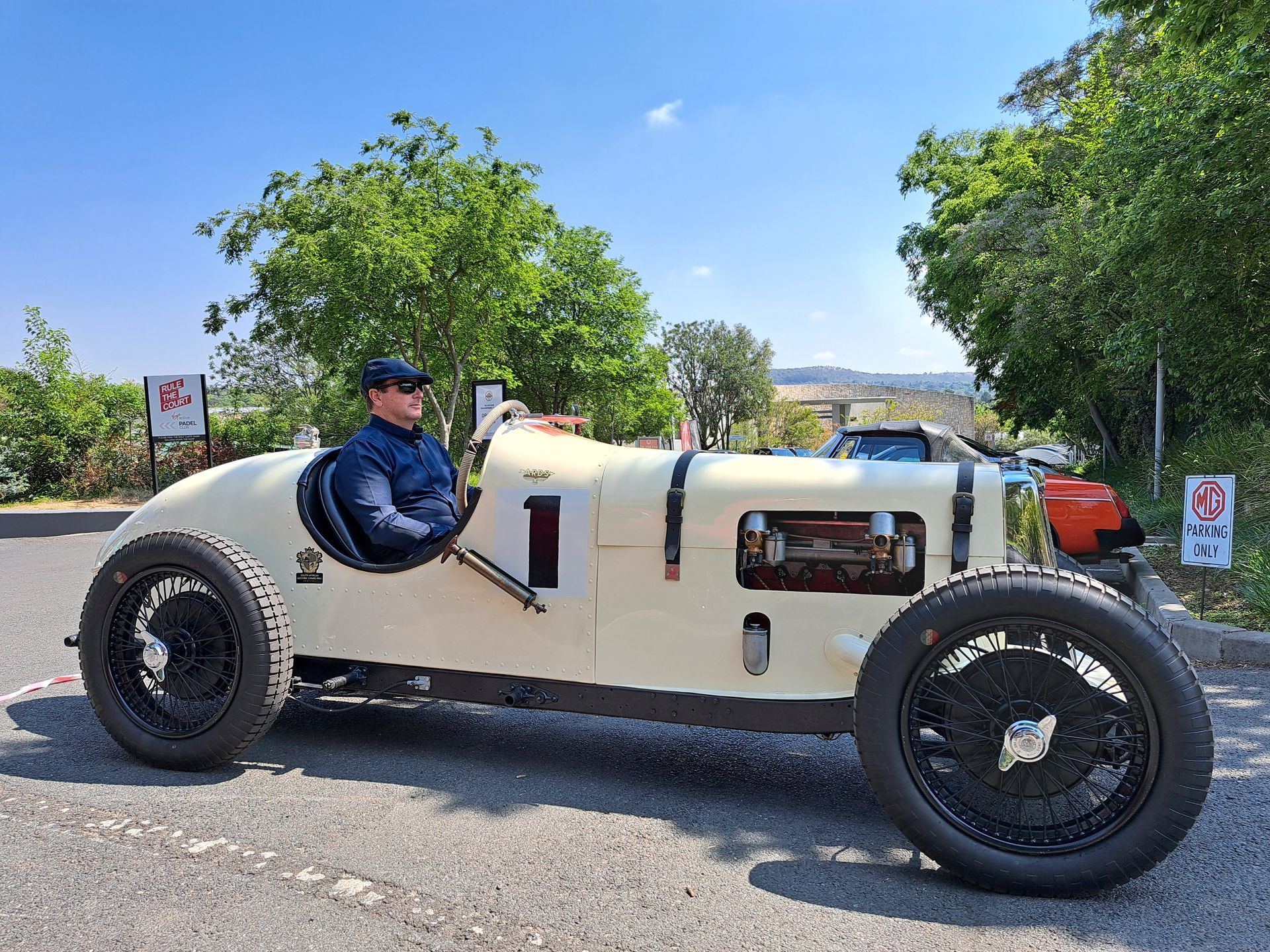 Vintage cream race car with a driver, parked outdoors on a sunny day.